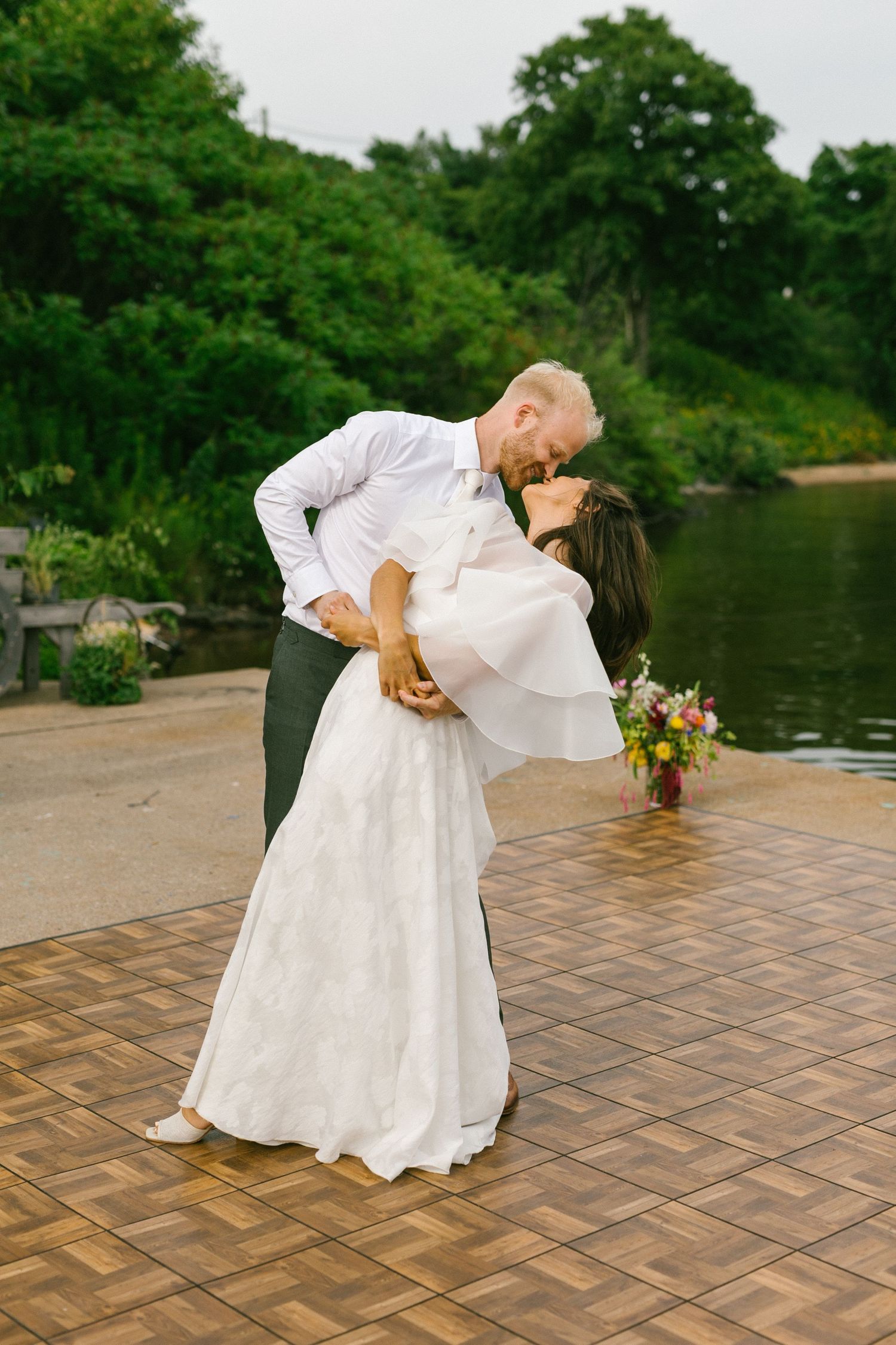 Wedding couple performs romantic dip dance move on outdoor brick patio near waterfront.