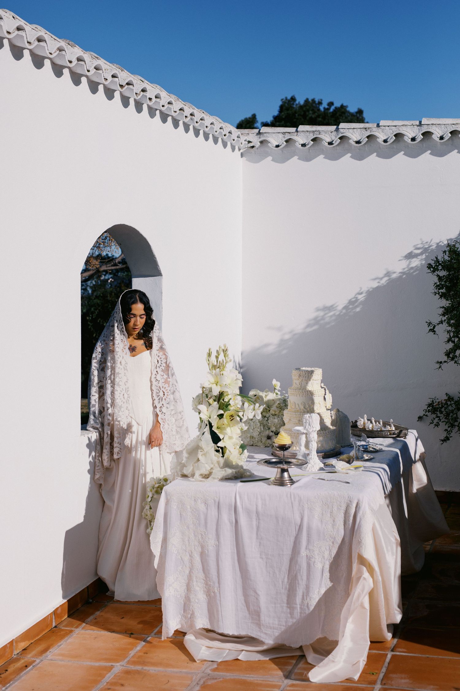 Bride in lace veil stands beside white wedding cake table at a whitewashed Mediterranean venue under blue sky.