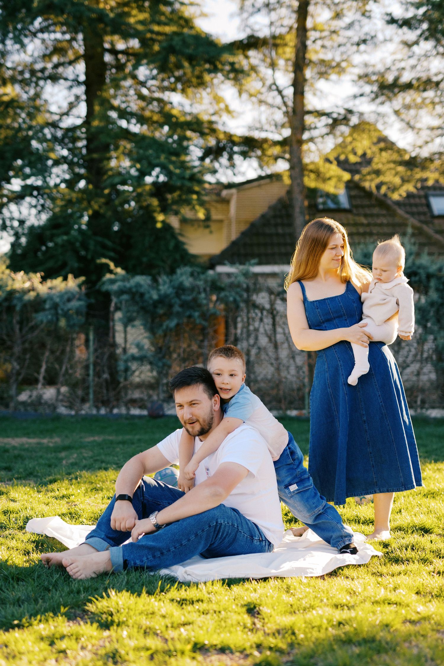 Happy family relaxing outdoors on a sunny day, with father sitting on grass, mother in blue dress holding baby.