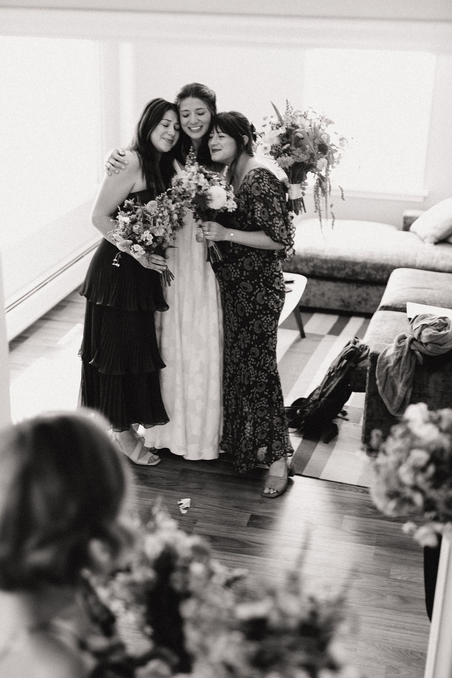 Three women in formal attire share an emotional embrace at an indoor celebration with floral decorations visible.