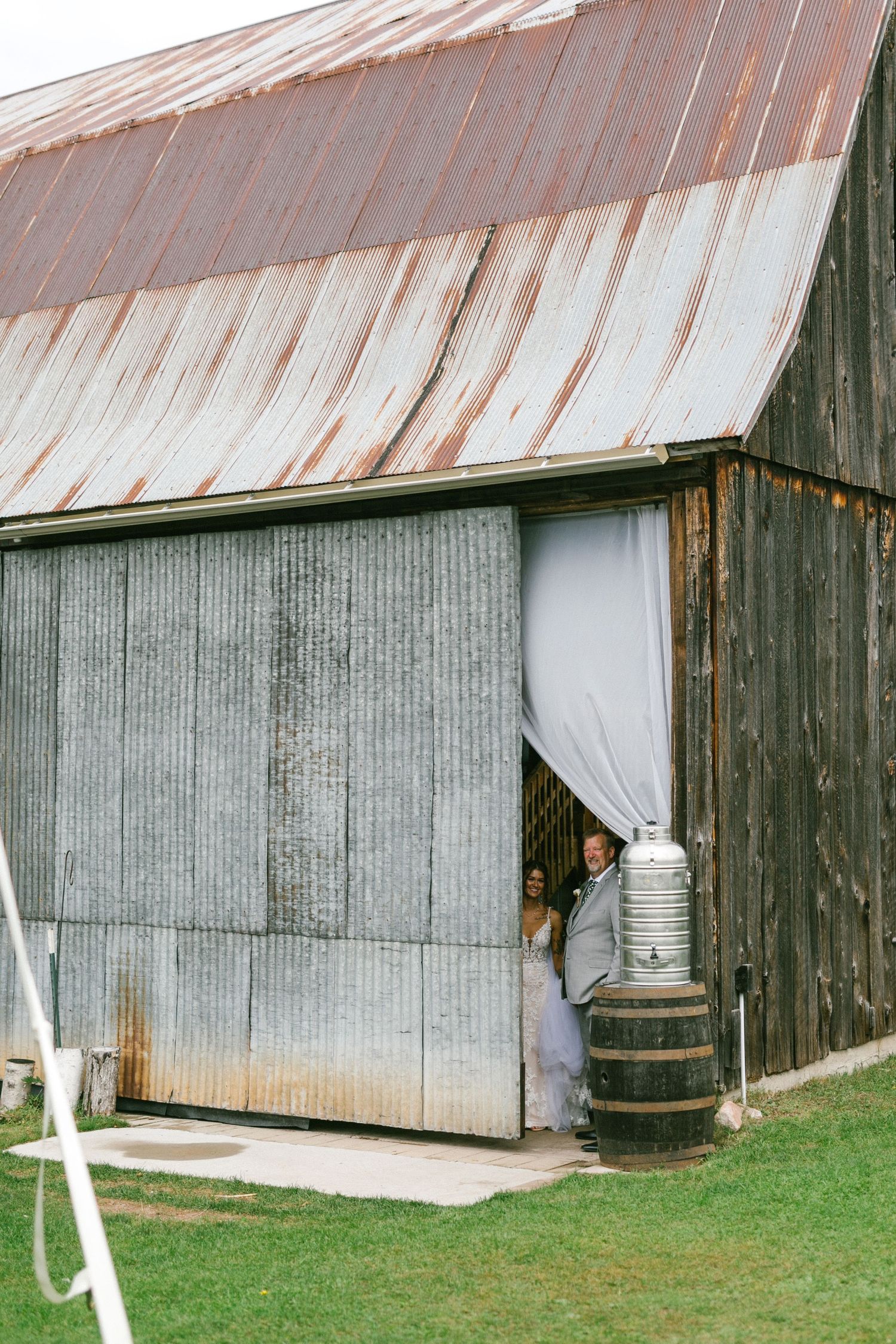 A bride and her father wait for the ceremony to begin in a rustic barn with corrugated metal walls and weathered roof in a rural setting.