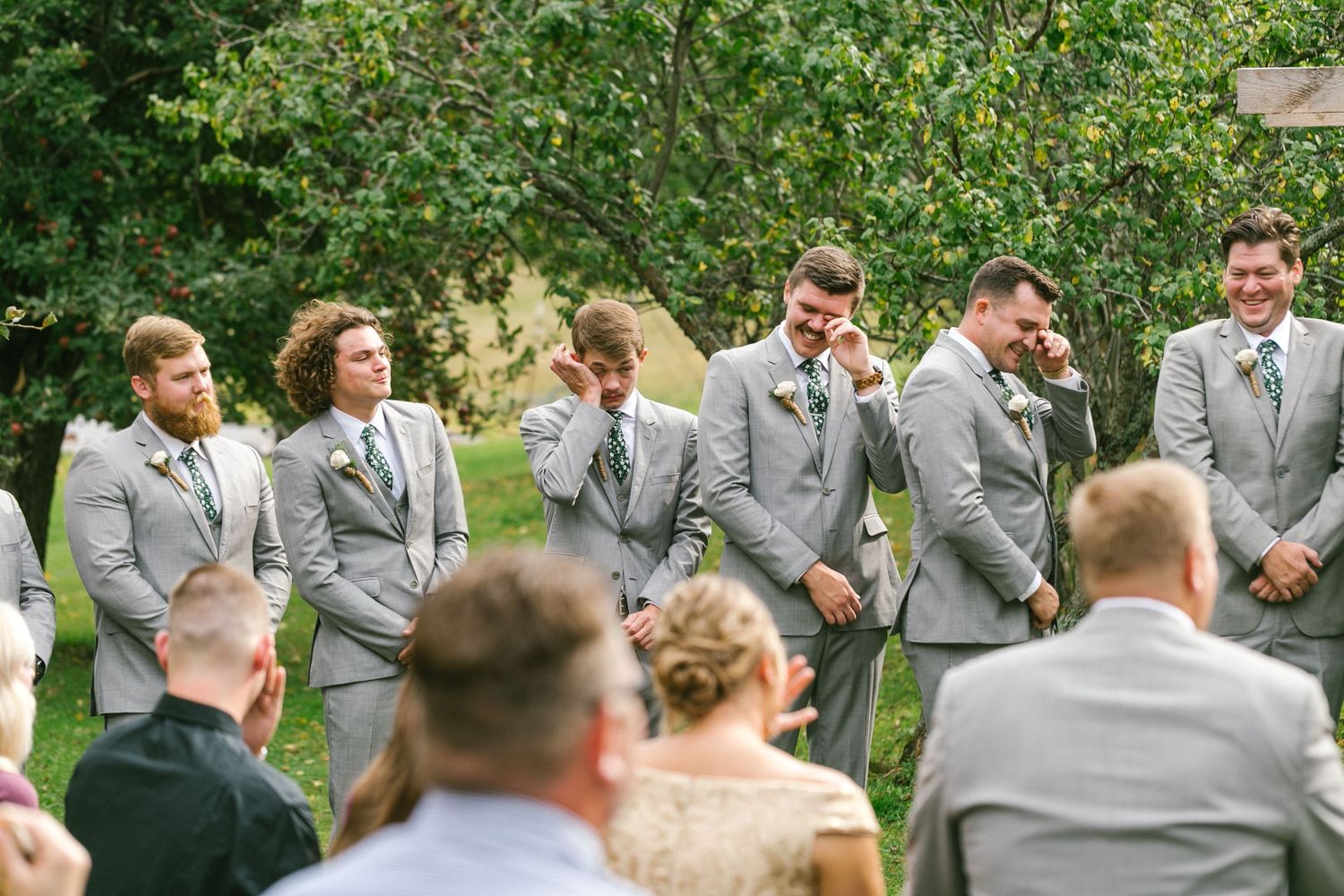 Wedding party members in matching gray suits share a lighthearted moment during an outdoor ceremony.