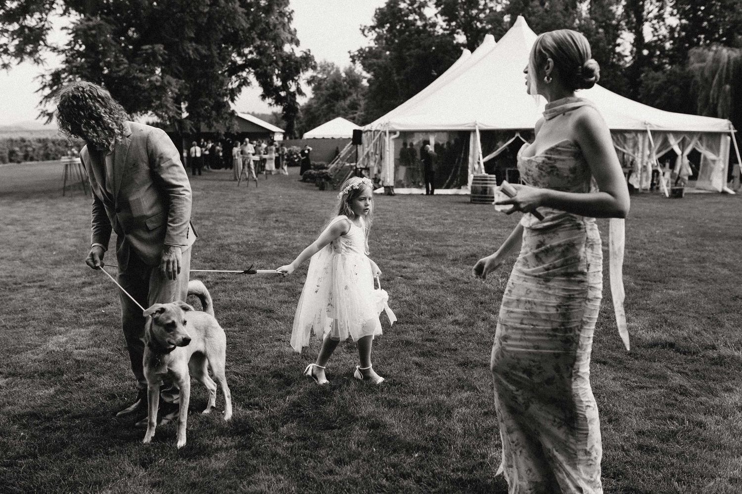 A black and white photo shows a girl tugs on a dog's leash as it wraps around another person.