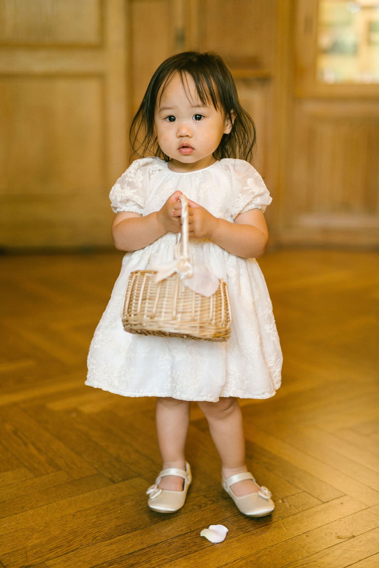 Adorable toddler flower girl in a white dress holds a wicker basket on a wooden floor.