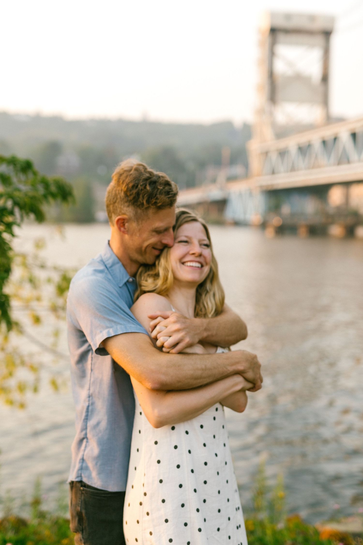 Couple embracing and laughing together by the river with a large bridge in the background.