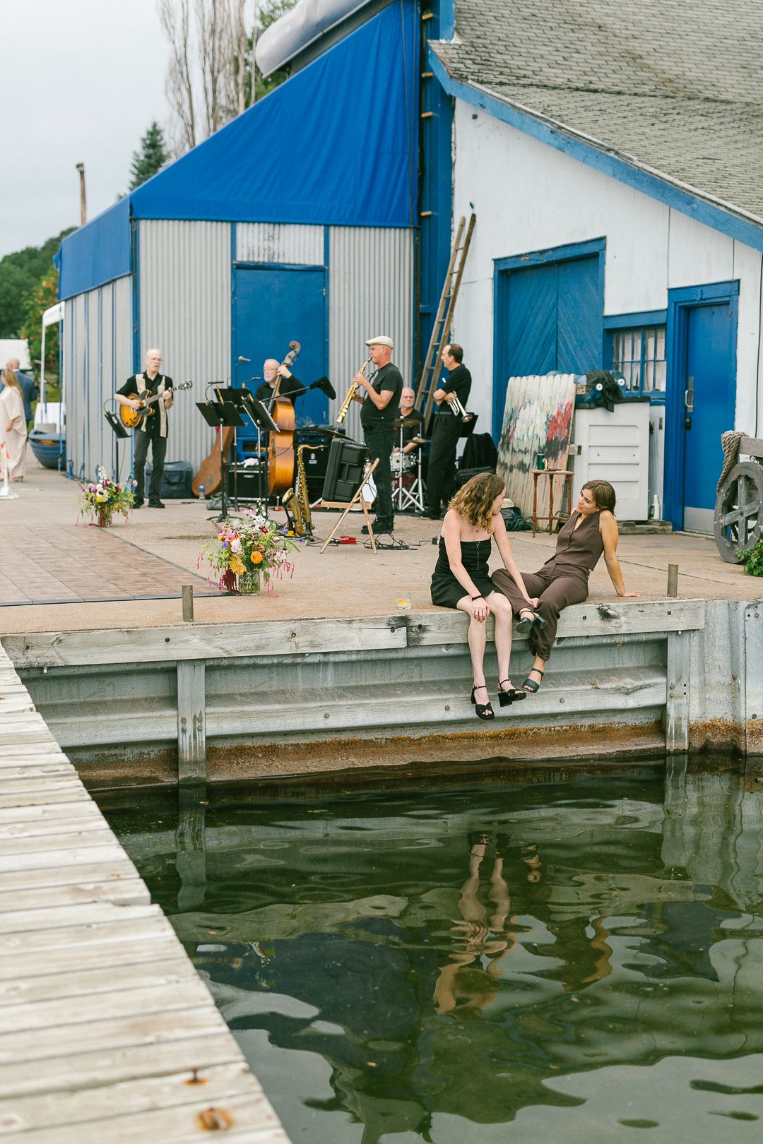 Wedding party members sitting along a wooden dock by a blue boathouse.