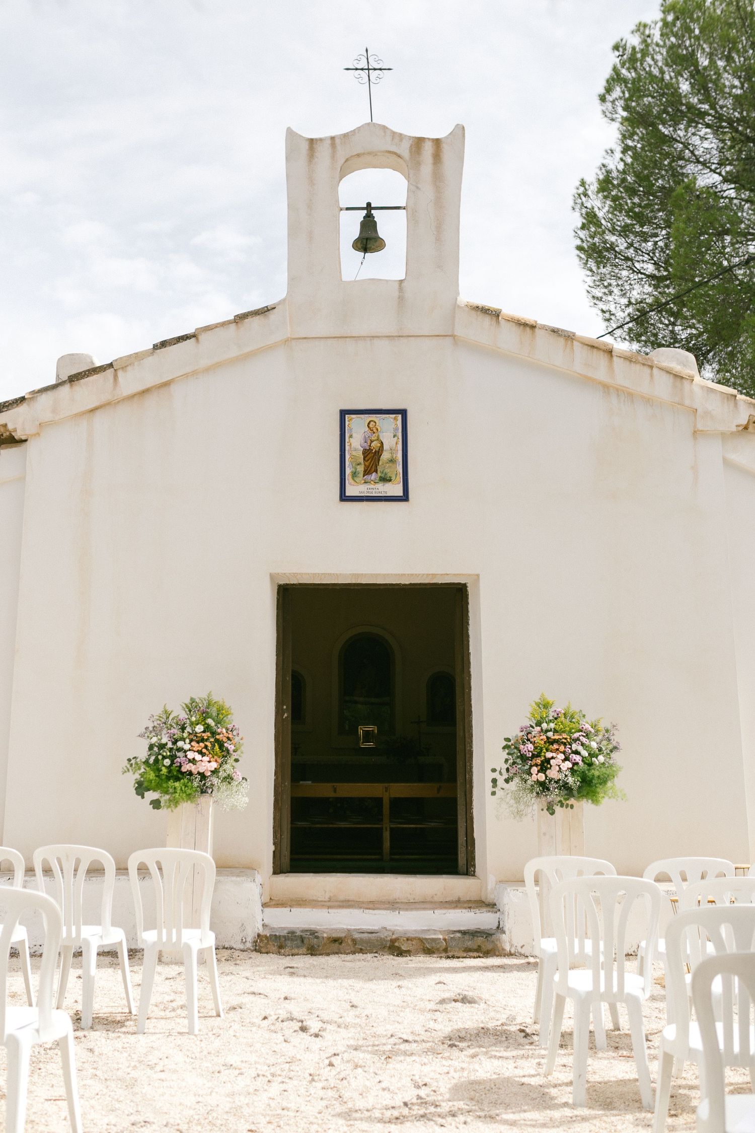 Small white chapel in Spain with bell tower and hanging flower baskets arranged with white chairs outside for a wedding ceremony.