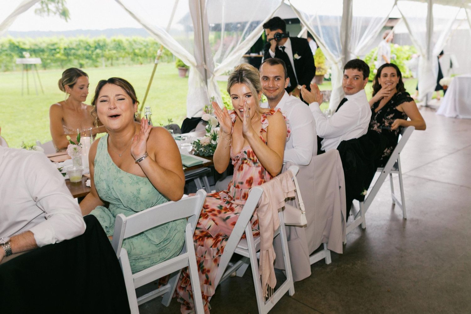 Wedding guests sharing joyful laughter under a white tent during an outdoor reception celebration.