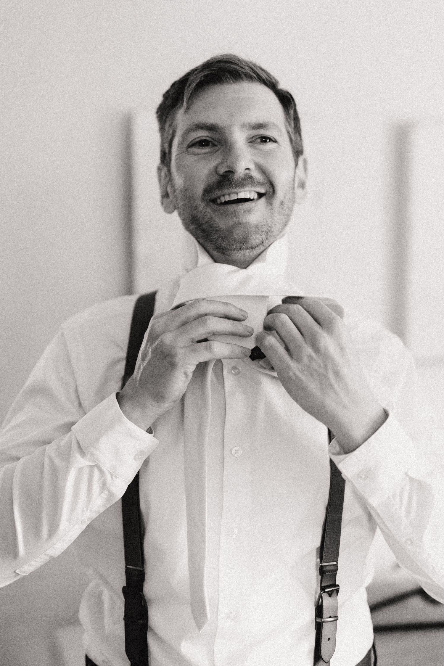 Black and white photo of someone adjusting their bowtie while wearing a white dress shirt and suspenders with a joyful smile.