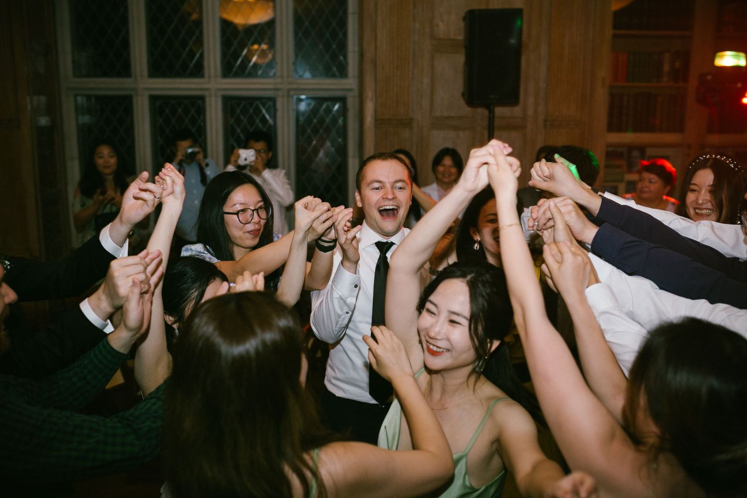 Joyful wedding reception dance floor with guests raising hands together in celebration at an elegant venue.