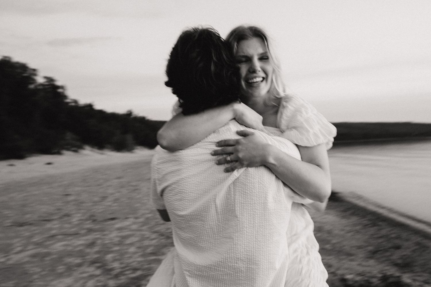 Black and white photo of two people embracing on a beach, one laughing joyfully, wearing white outfits.