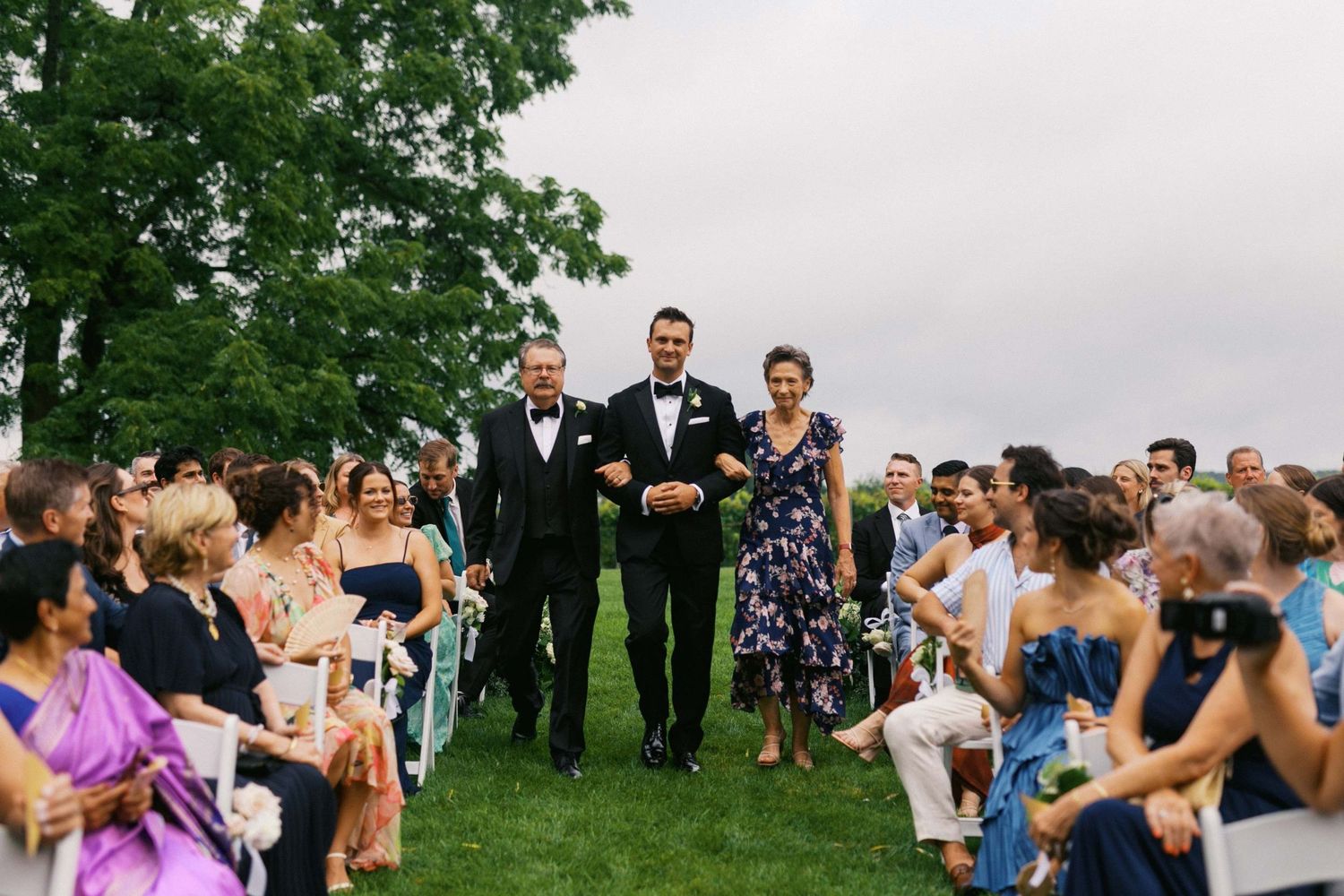 Wedding guests stand outdoors on a grassy lawn watching a special moment during a summer celebration.