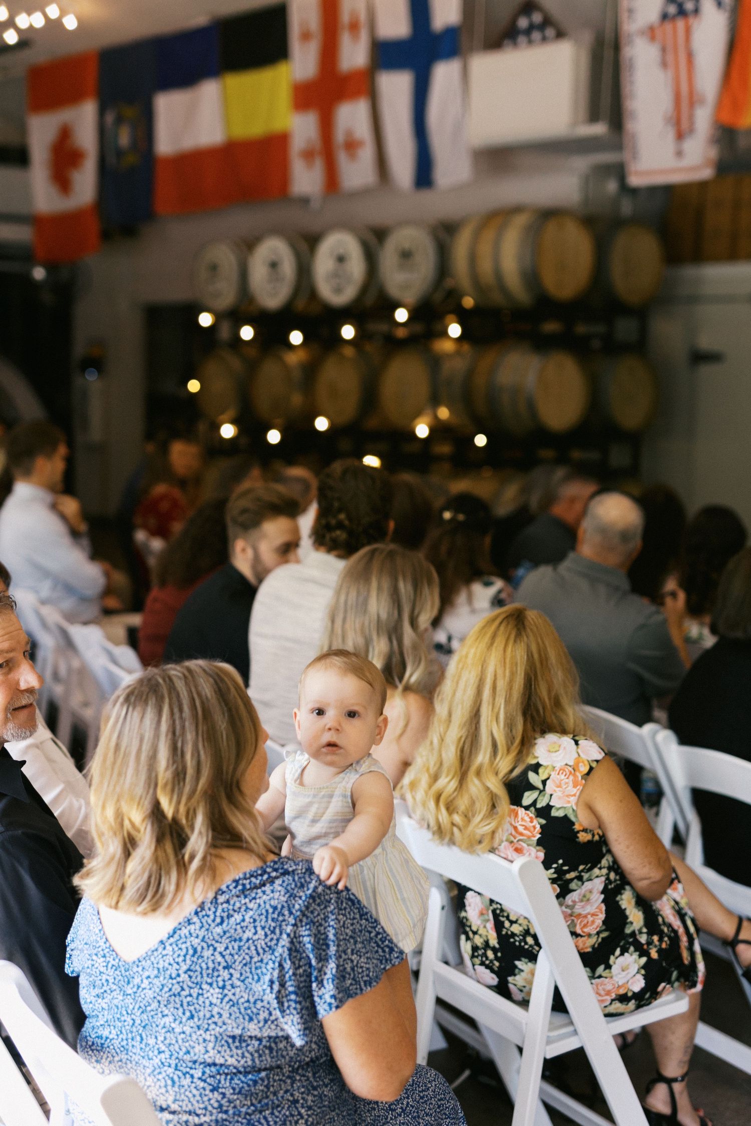 Wedding guests seated in white chairs at a rustic venue with wine barrels and string lights in the background.