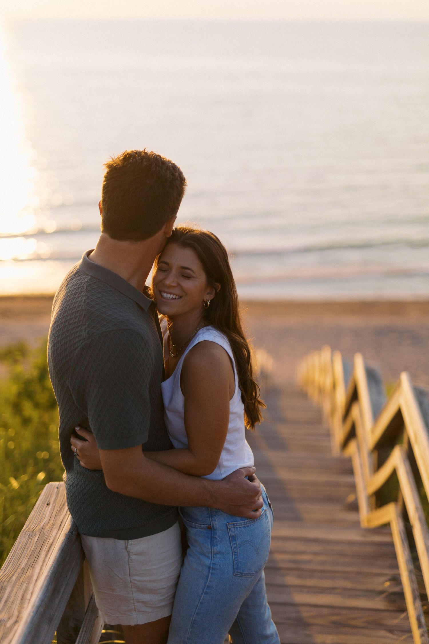 A couple embraces on a wooden boardwalk at sunset with the ocean glowing in the background.