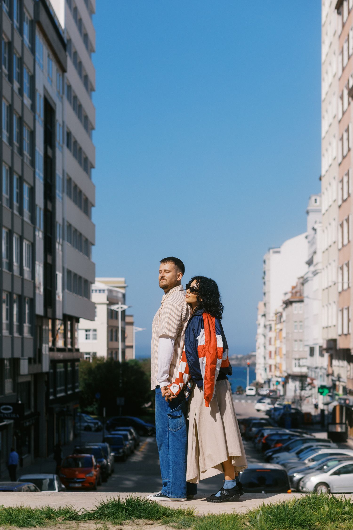 A couple stands back-to-back on a sunny urban street flanked by modern buildings with ocean views in the distance.