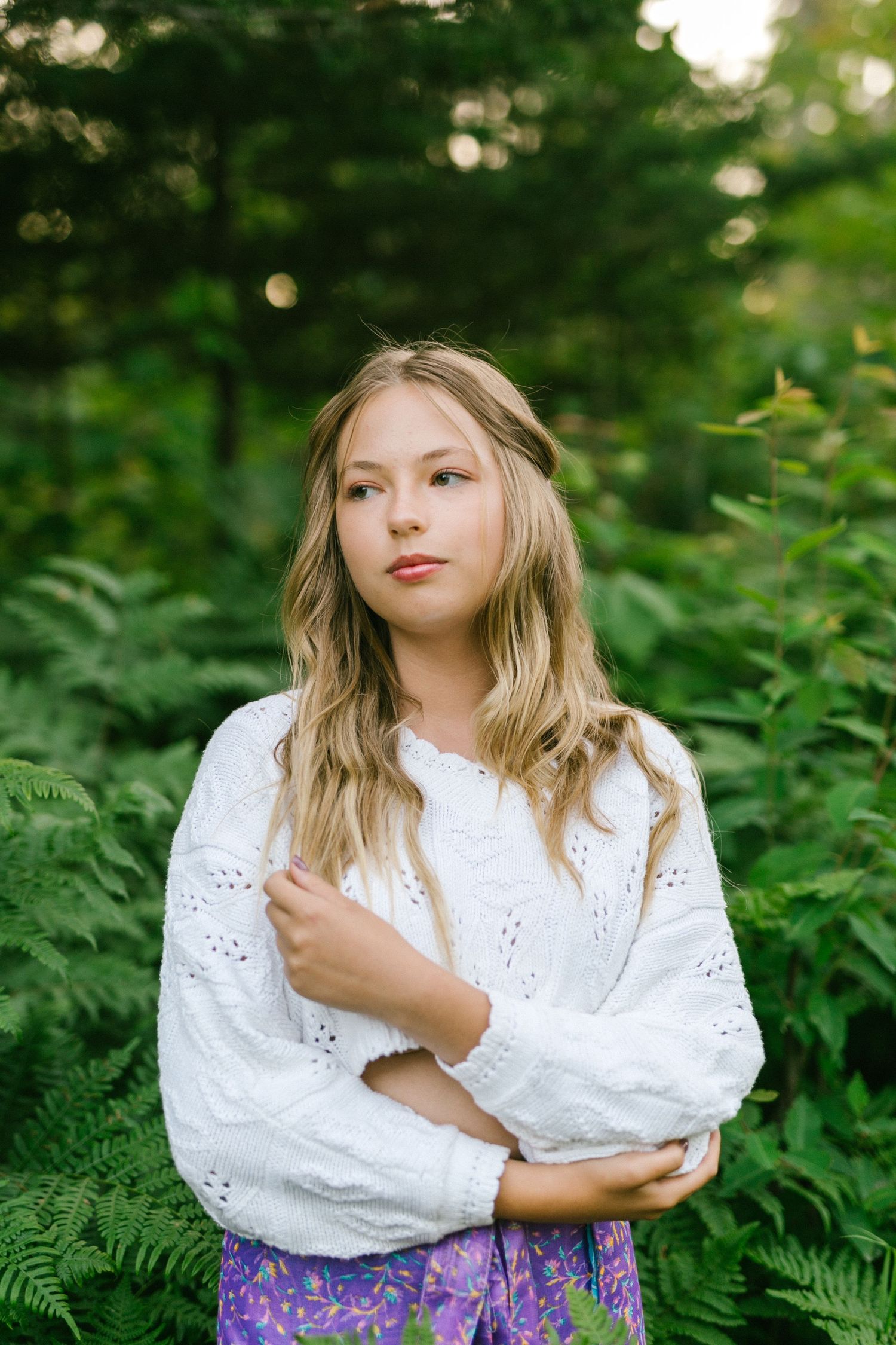 Blonde teenage girl in white eyelet sweater and purple skirt poses thoughtfully among lush green ferns outdoors.
