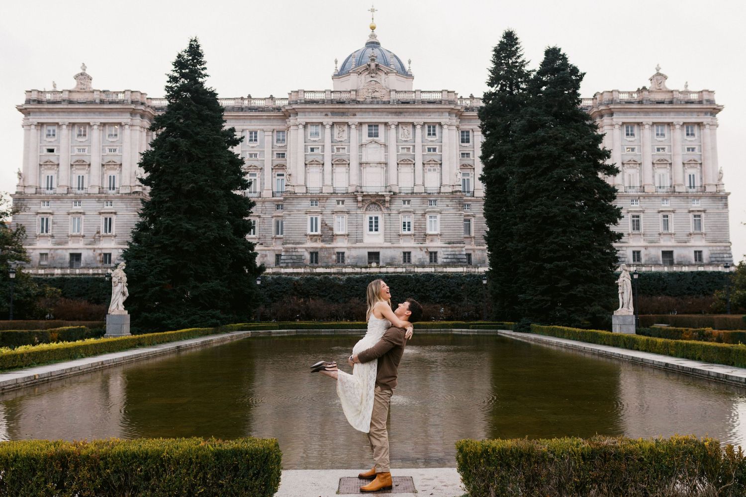 Romantic couple dancing in front of the Royal Palace of Madrid near a reflecting pool.
