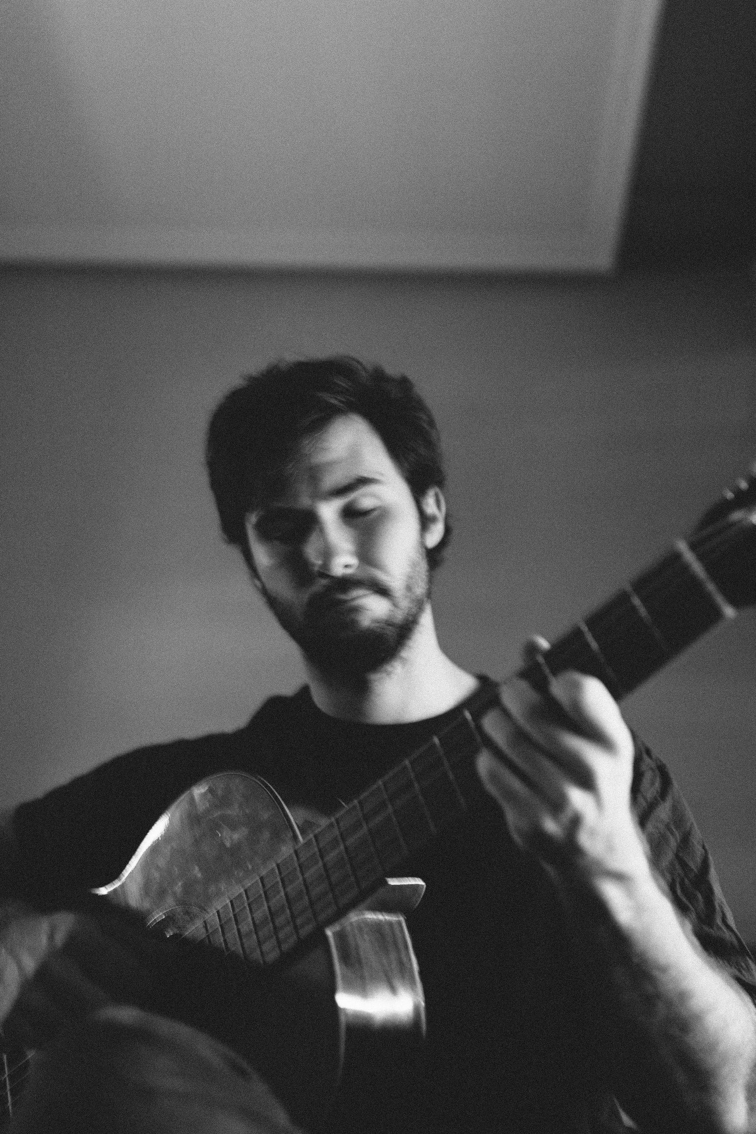 Black and white portrait of a bearded man playing acoustic guitar indoors.