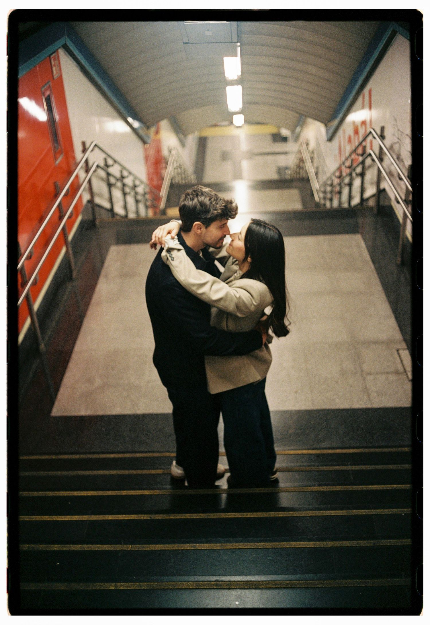 Couple embracing and kissing on subway station stairs with red train carriages in background.