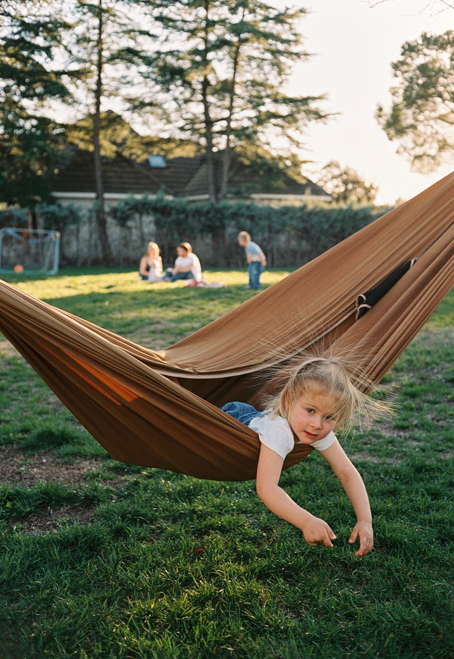 Young blonde girl playing in a brown hammock in a sunny backyard with family relaxing in the background.