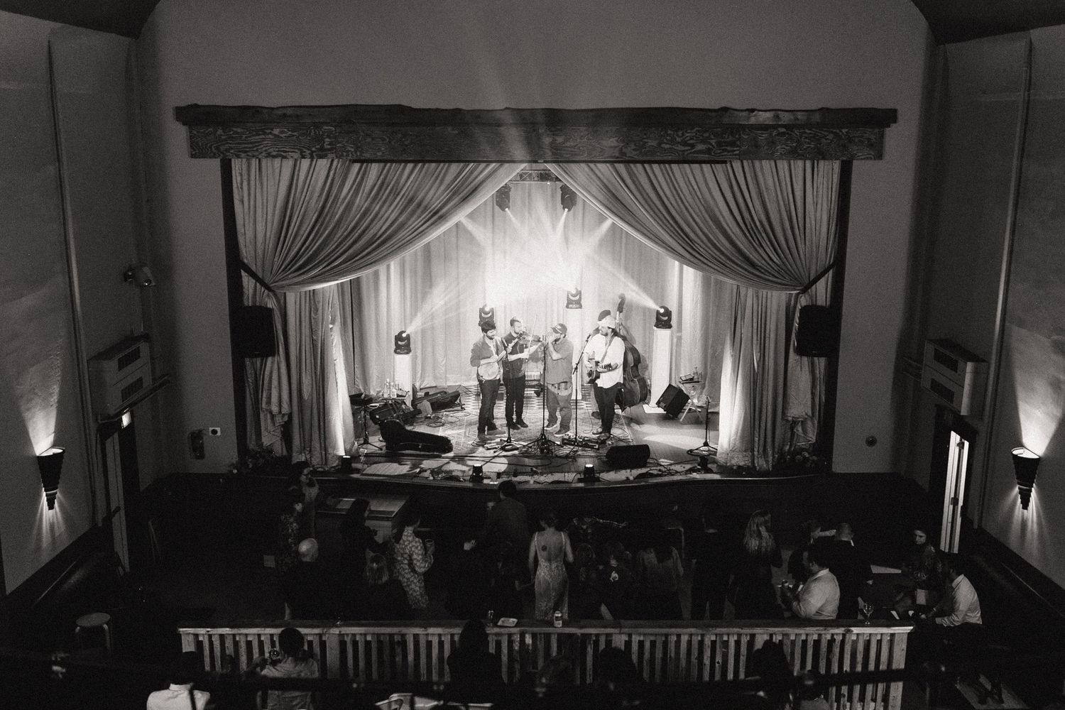 High-contrast black and white image of a band performing on stage in an intimate theater venue with dramatic curtains.