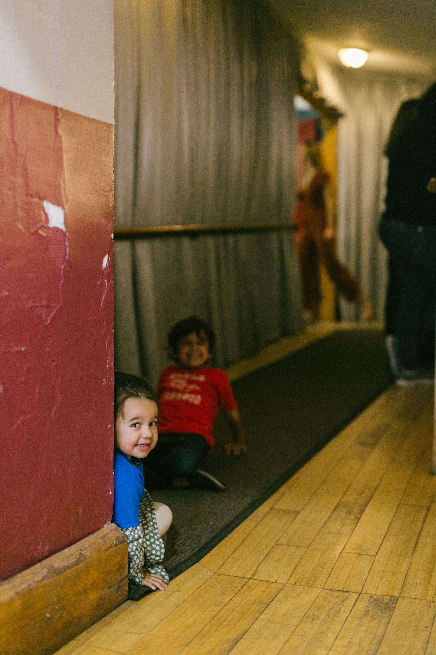 Children playing and hiding in a dimly lit hallway with wooden floors and red walls.
