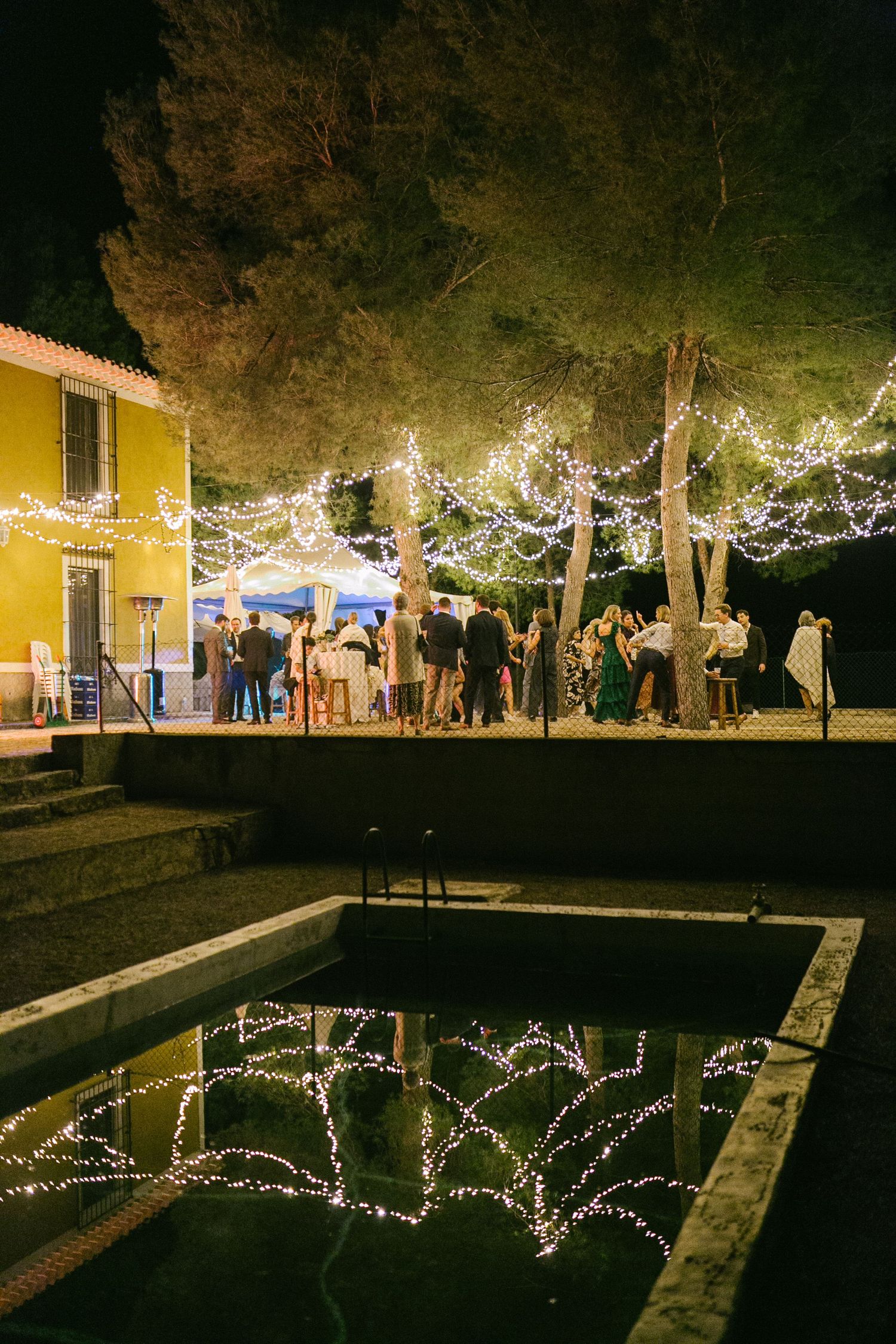 Evening party by a pool with string lights reflecting in the water and a yellow building in the background.