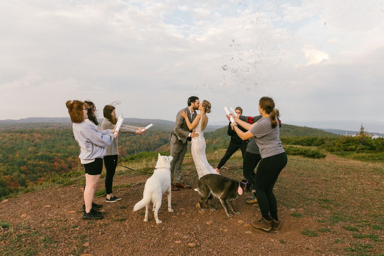 A joyful celebration takes place on a hilltop with a white dog joining the group against a scenic mountain backdrop.
