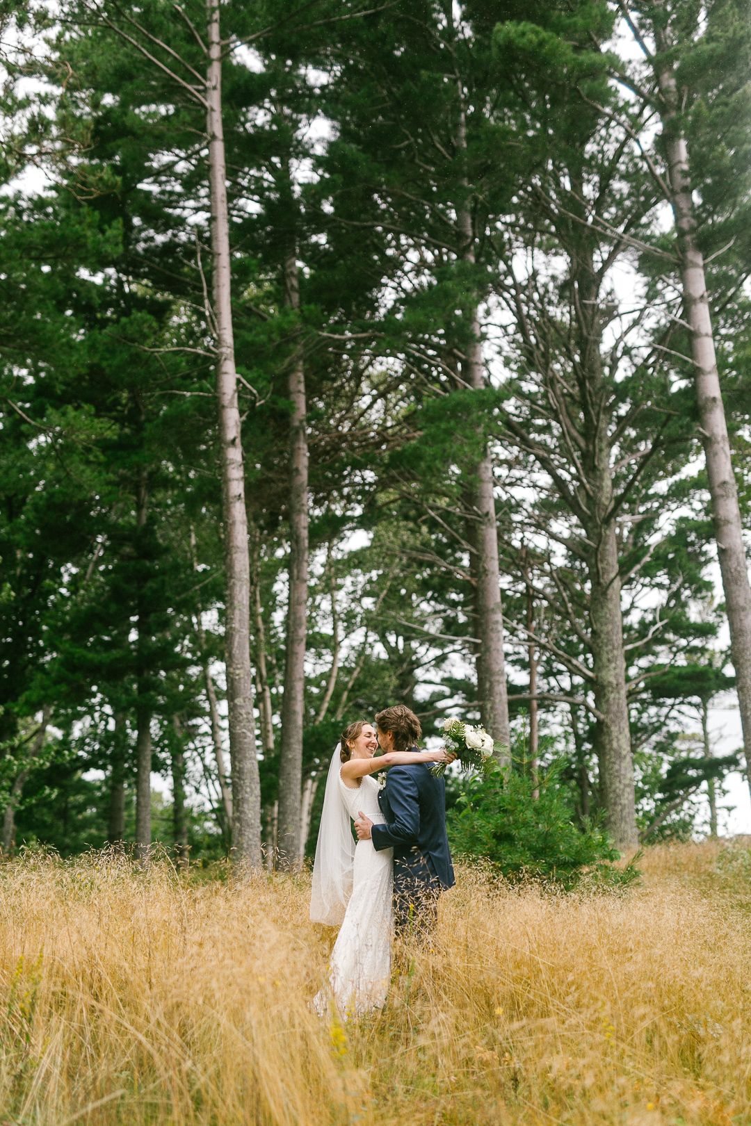 A couple stands among tall pine trees in a golden wheat field.