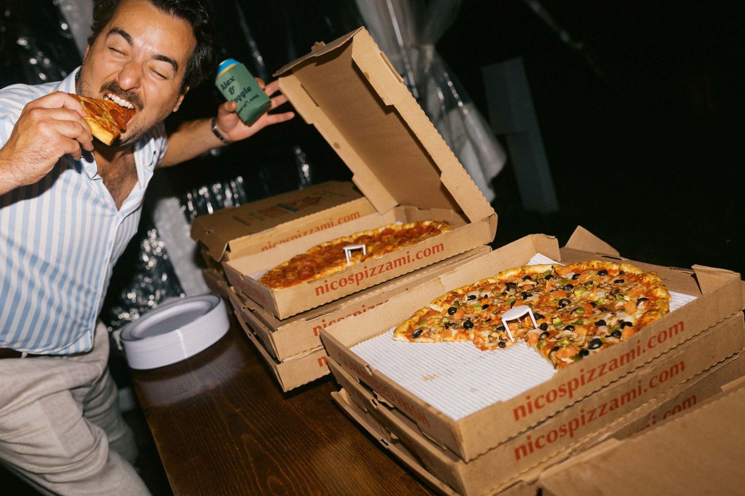 A person enjoying pizza and drinks with multiple pizza boxes stacked on a dark table.