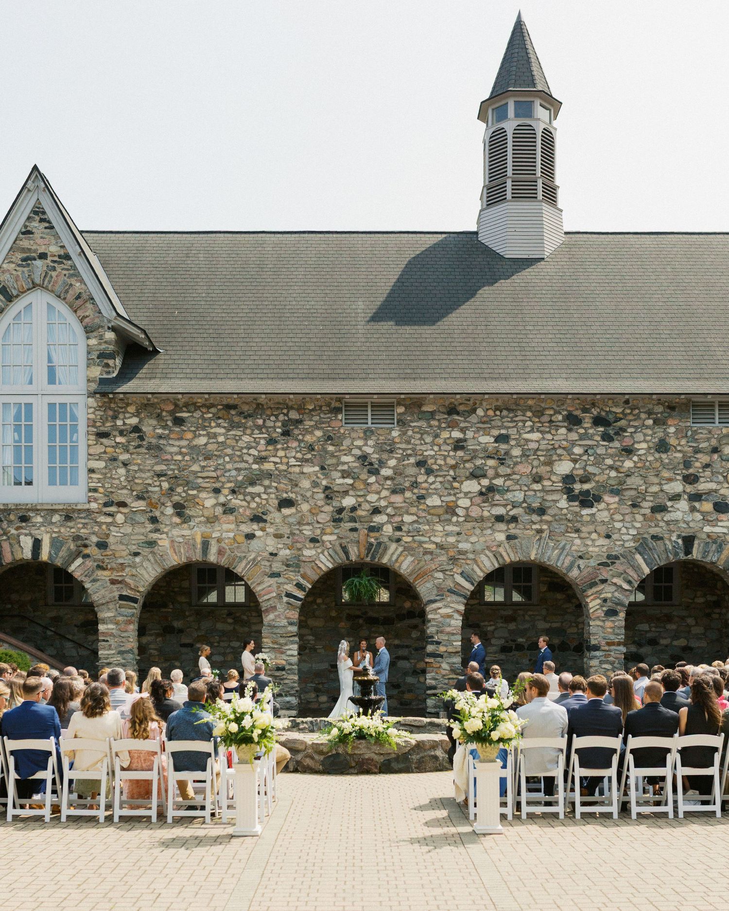 Outdoor wedding ceremony setup with stone chapel backdrop, white chairs, and floral arrangements.