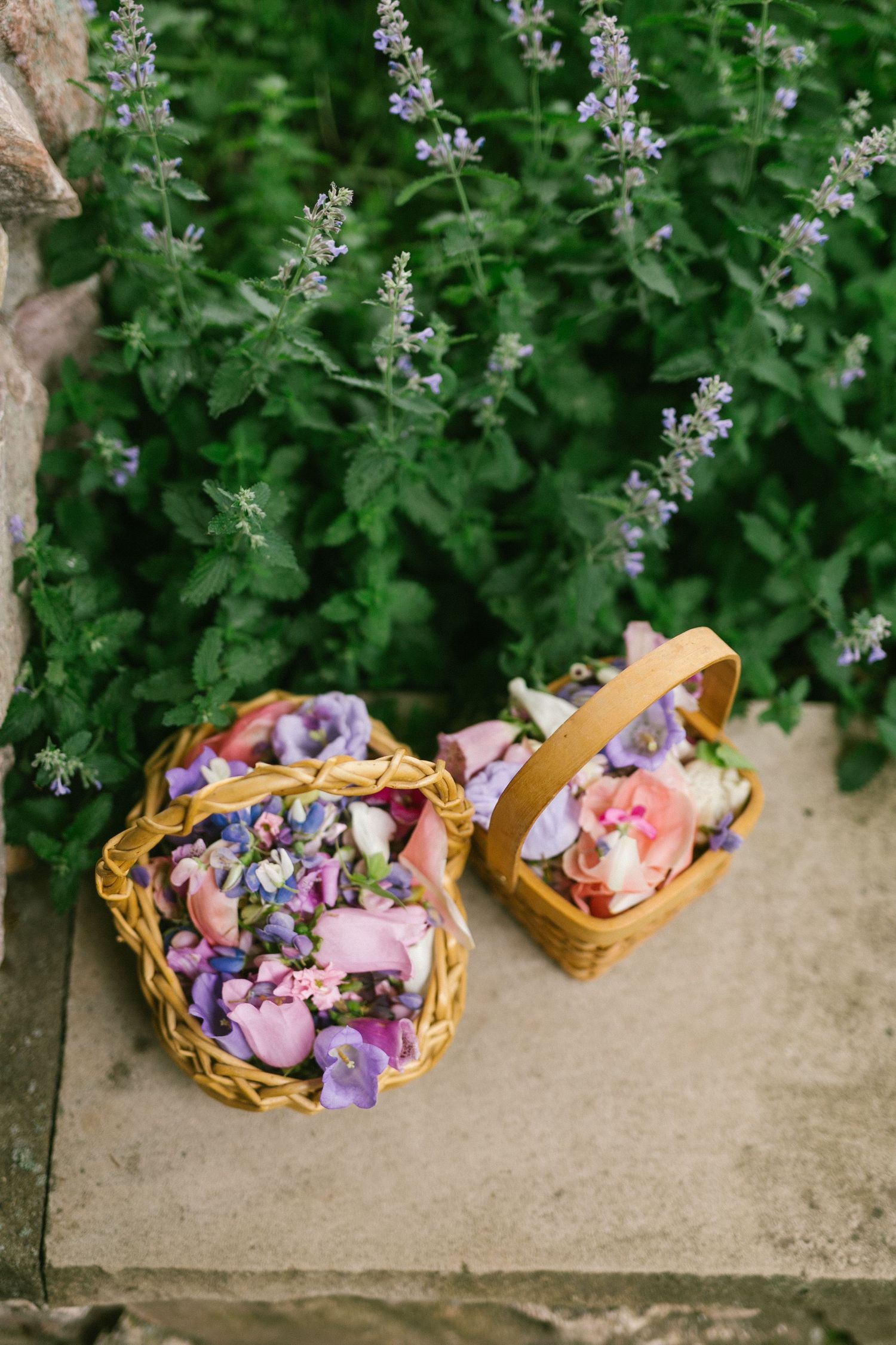 Two woven flower girl baskets filled with pink and purple petals sit on stone steps against flowering greenery.