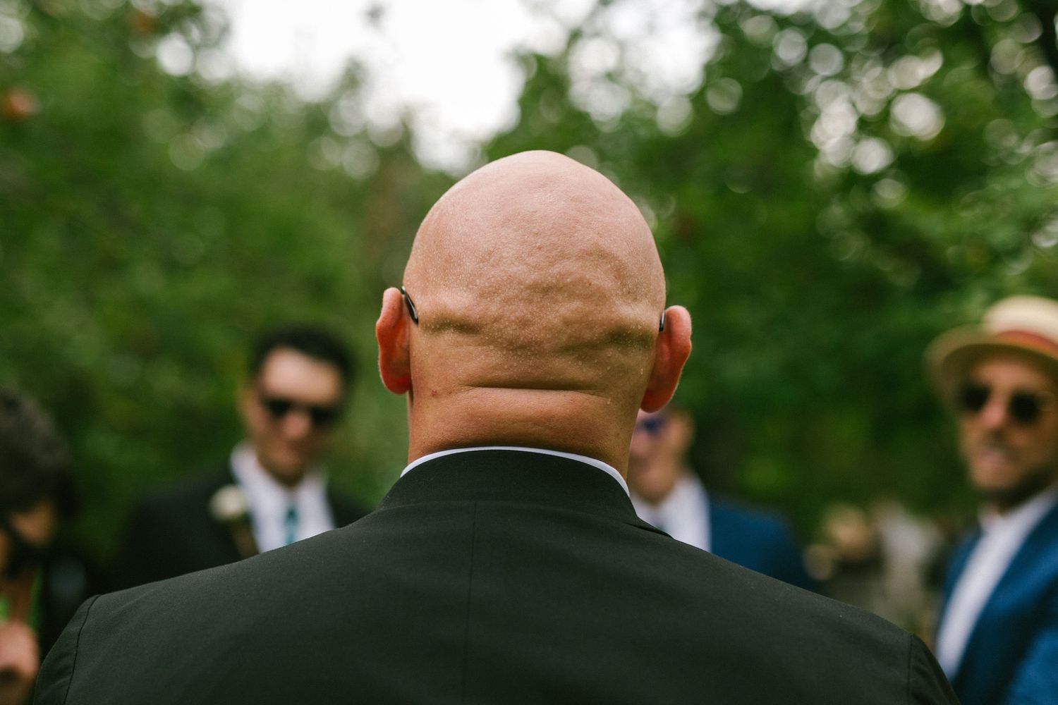 A bald person in a dark suit jacket is viewed from behind at an outdoor event, with other guests visible in the background.