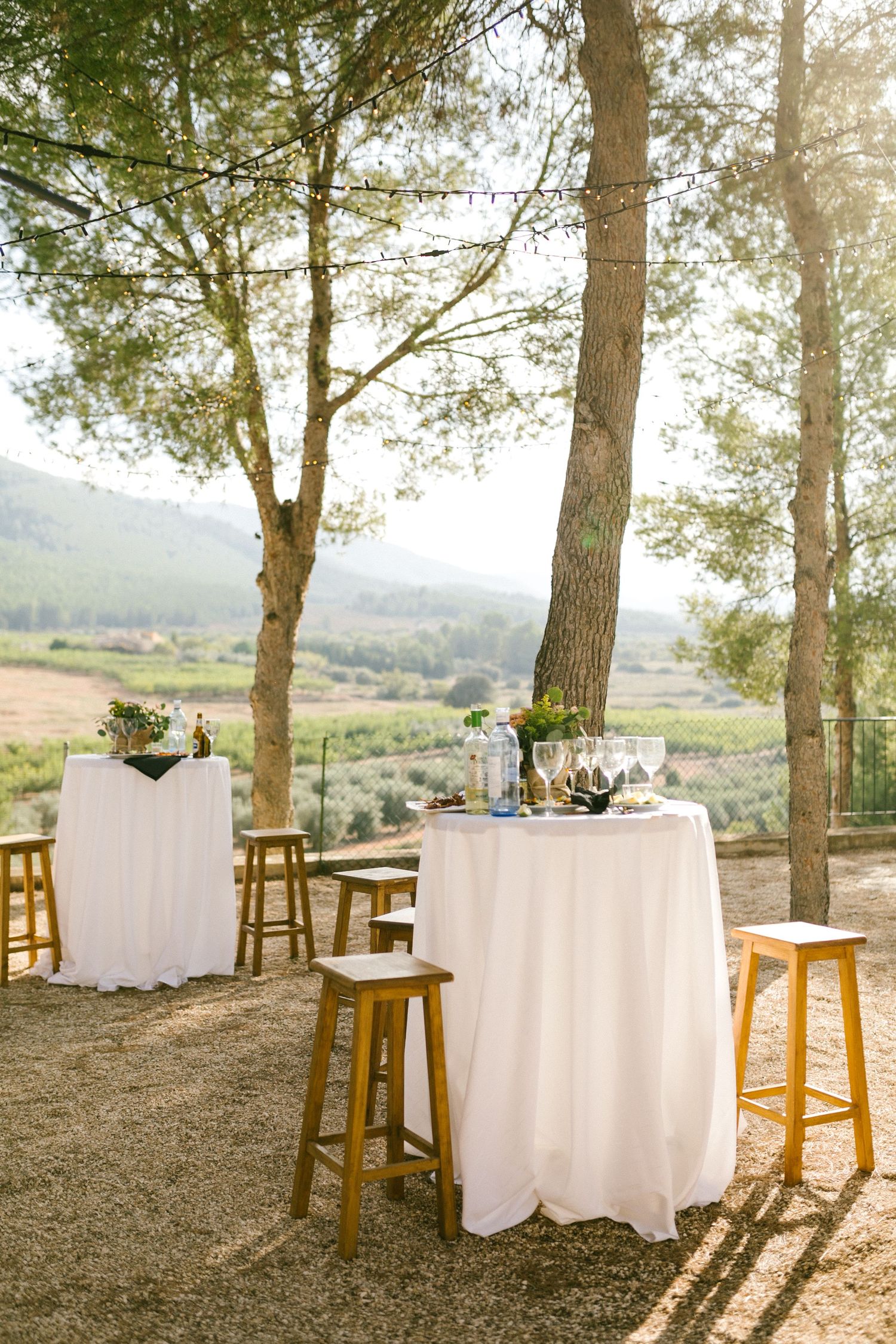 Elegant outdoor event setup with cocktail tables draped in white linens and wooden stools under tall pine trees at sunset.