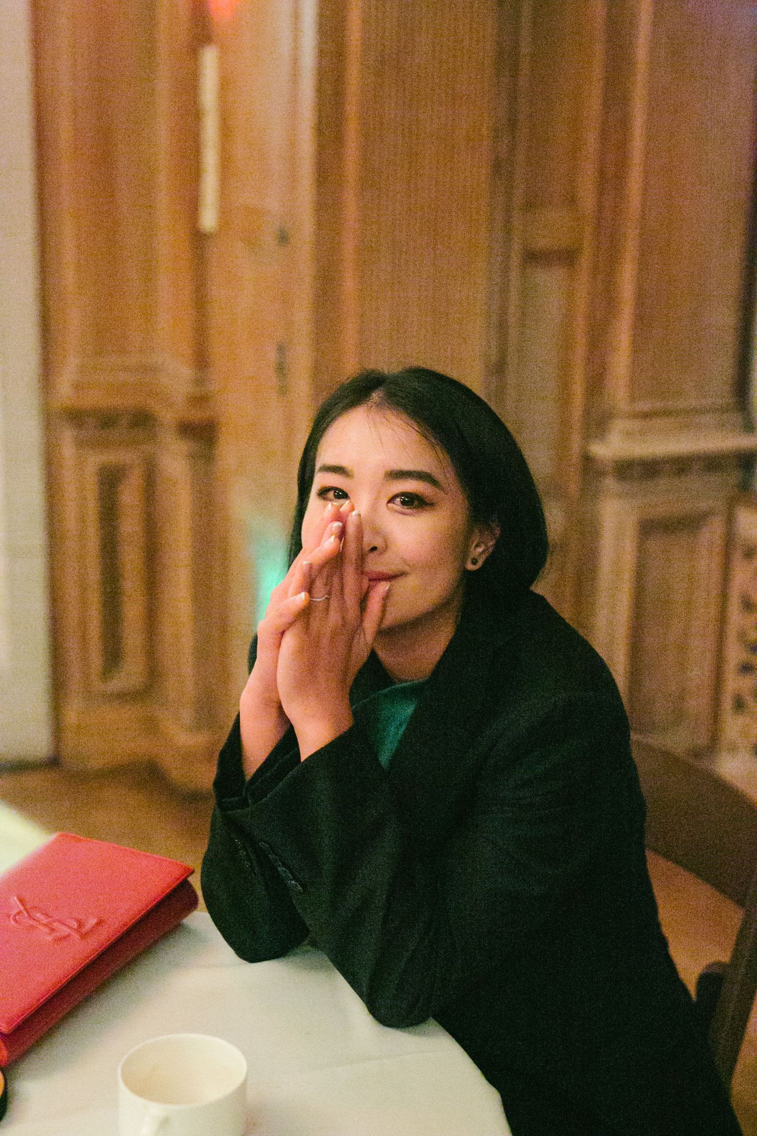 Young Asian woman sitting at a table with hands near her face, smiling warmly in a cozy indoor setting.