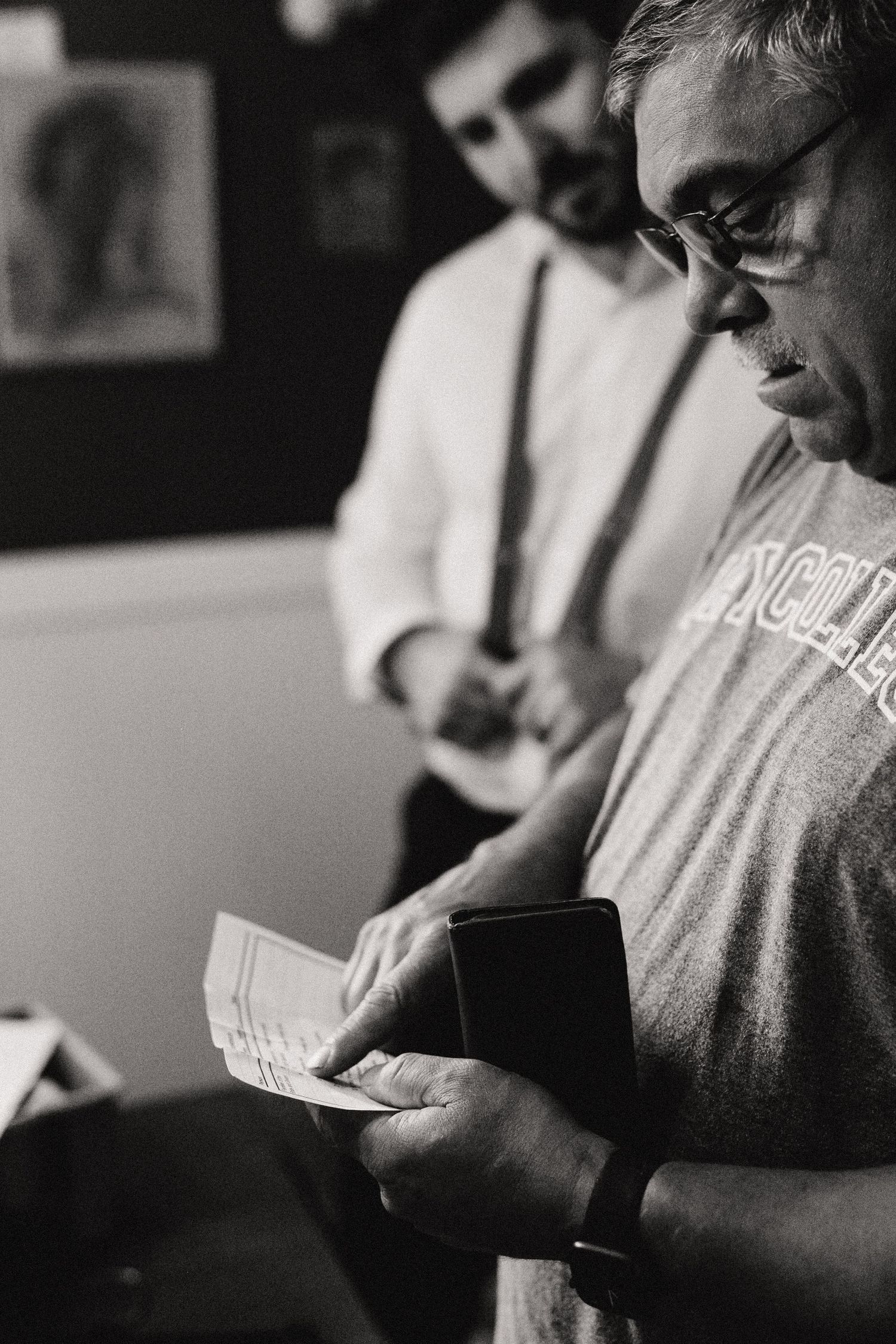 A black and white photograph shows a person in glasses reading papers while another figure stands in the background.