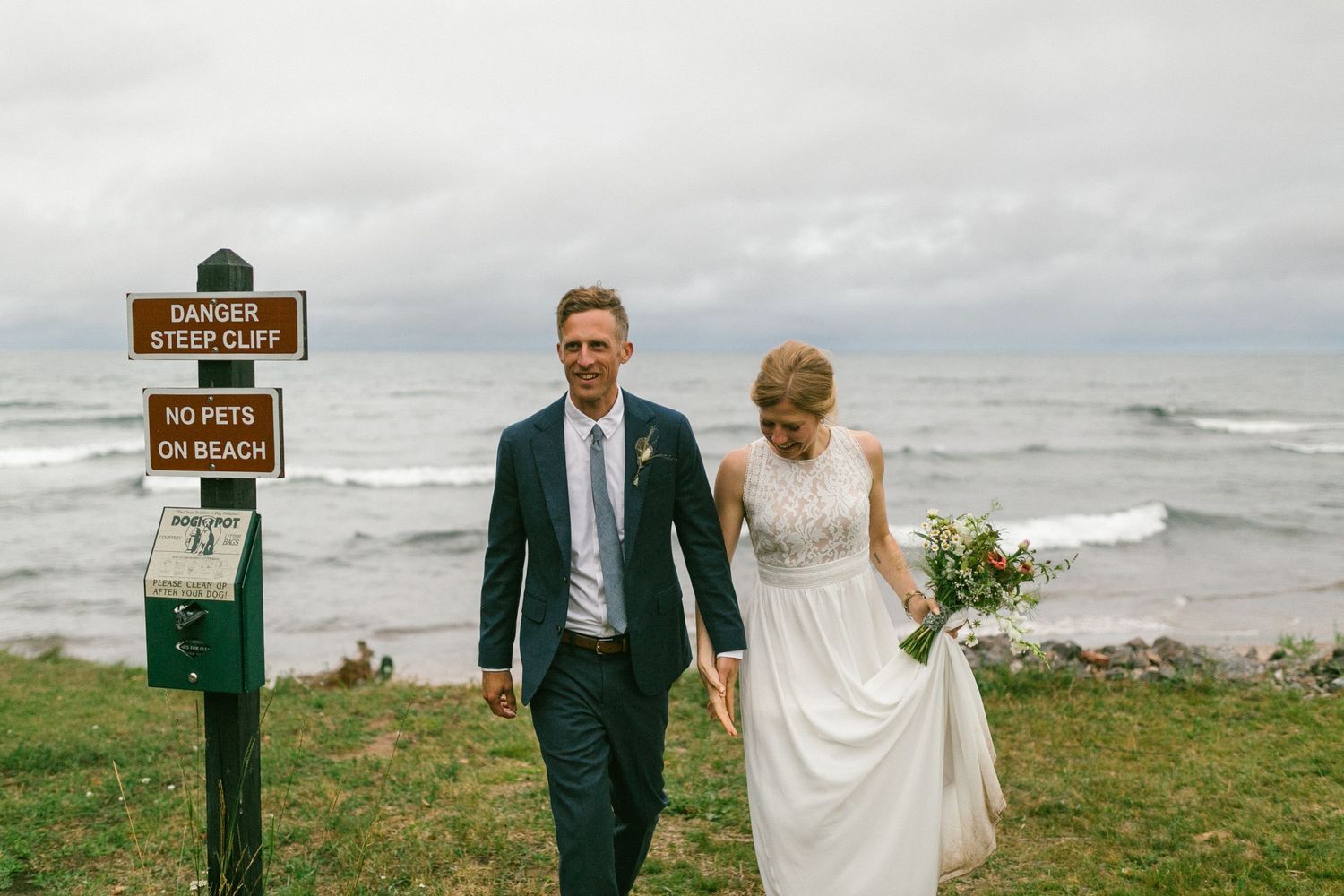 Two people stand near a wooden trail marker sign on a coastal bluff overlooking Lake Superior.