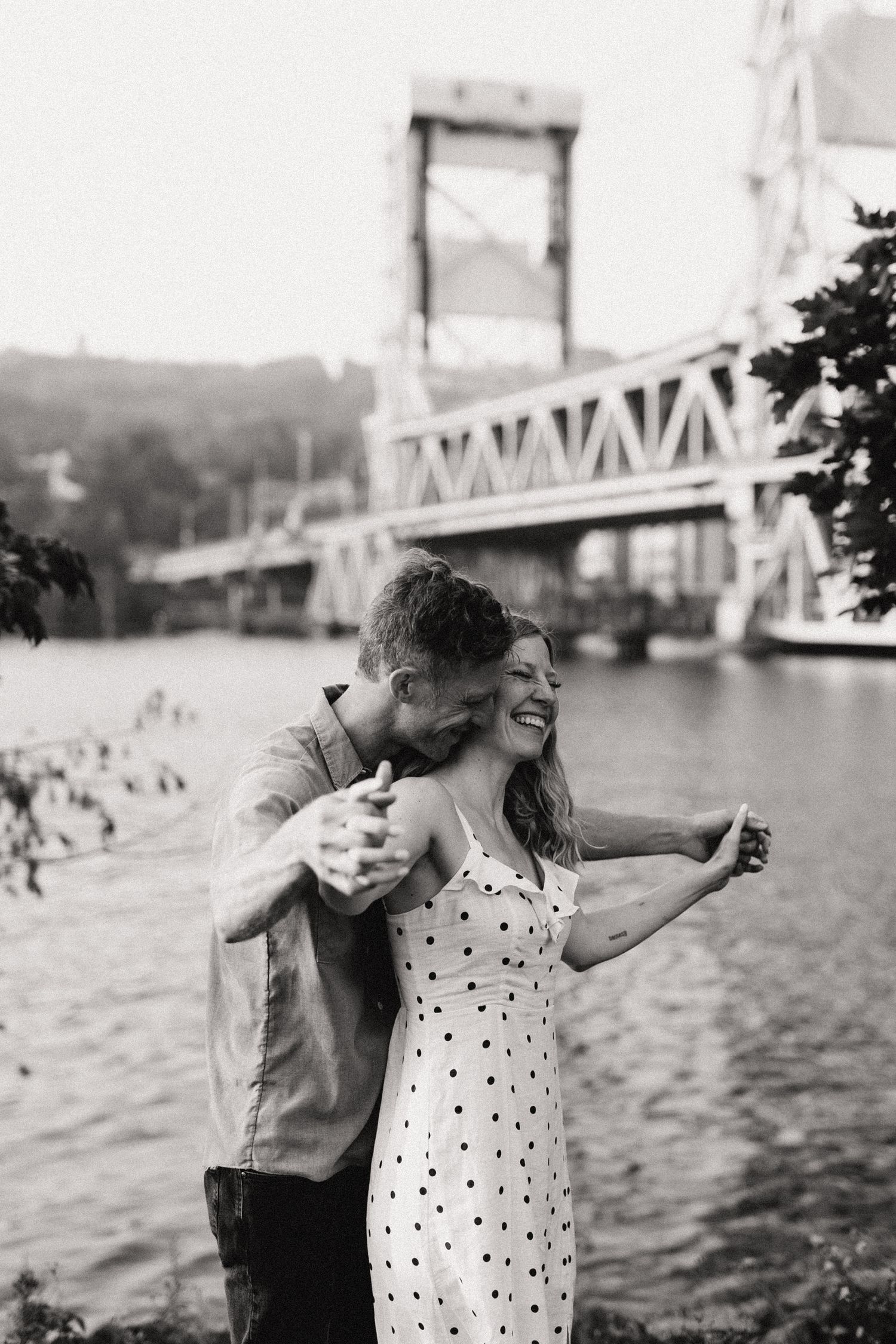Black and white photo of a couple laughing together by a bridge over water.