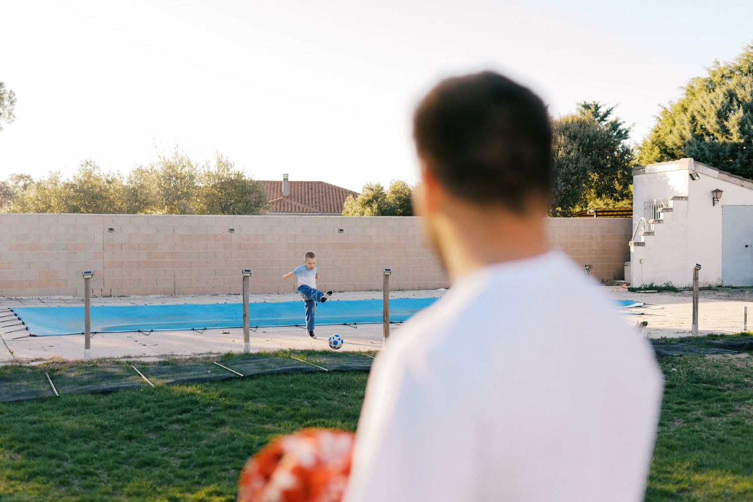 Two people playing soccer near a backyard swimming pool on a sunny day.