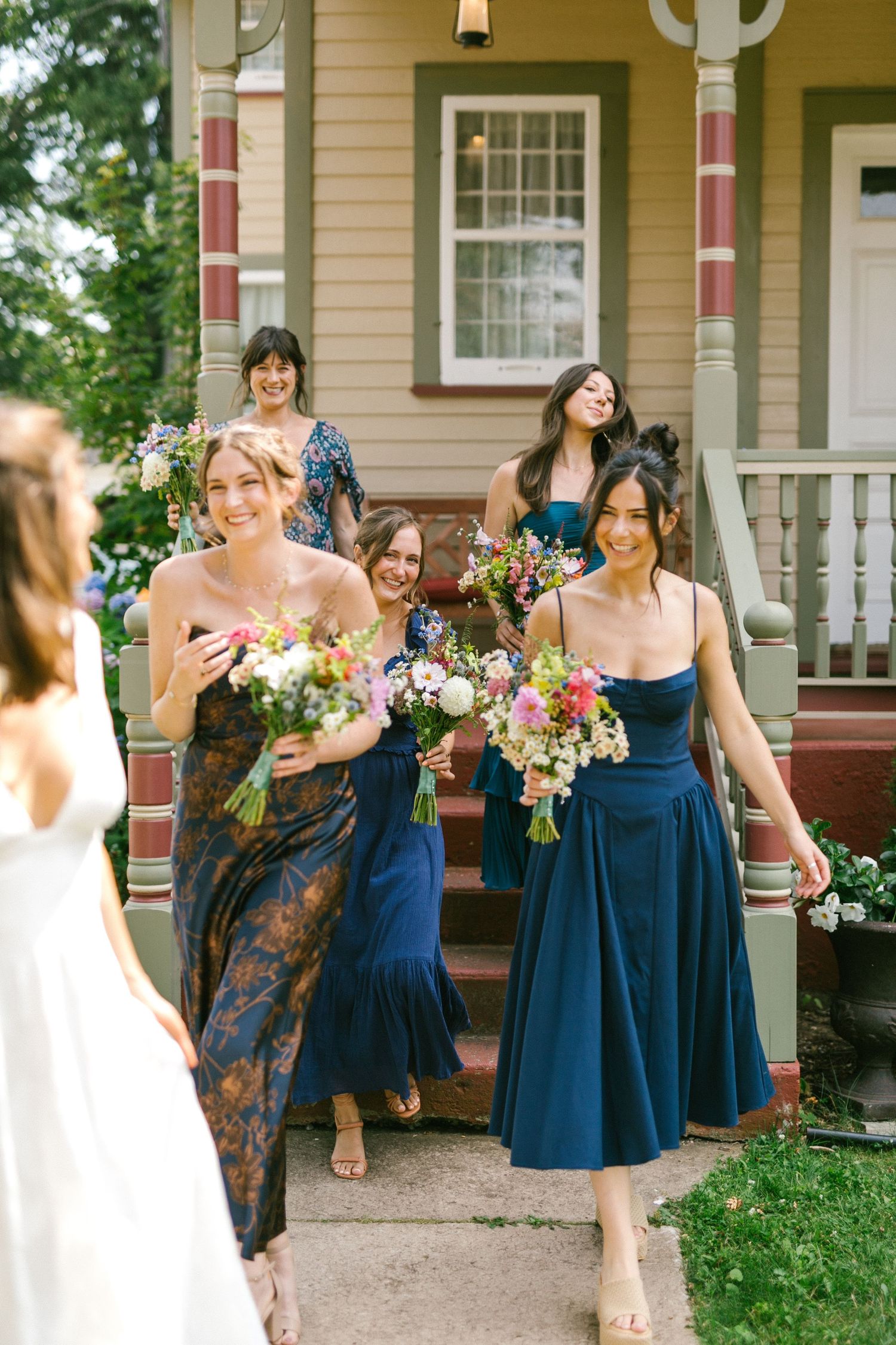 A wedding party in navy blue bridesmaid dresses gather outside a traditional house with colorful bouquets.