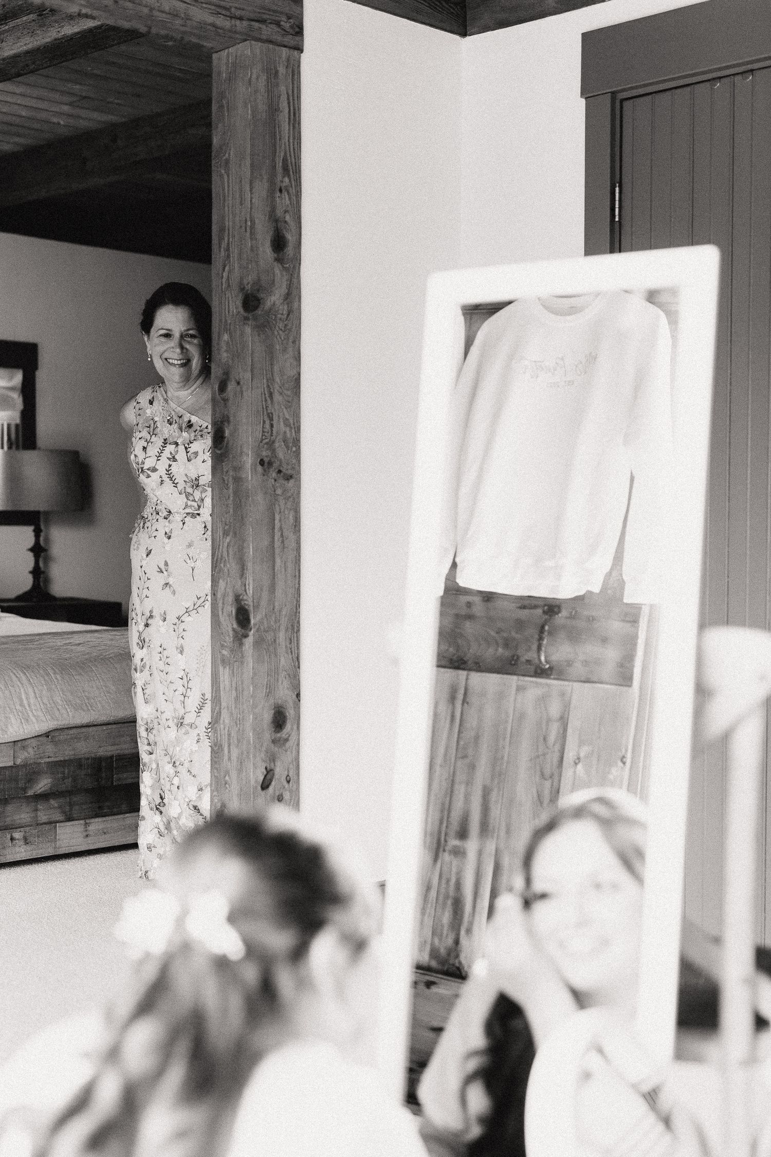 Black and white photo of a smiling woman in a floral dress standing in a doorway, with a mirror reflection in the foreground.