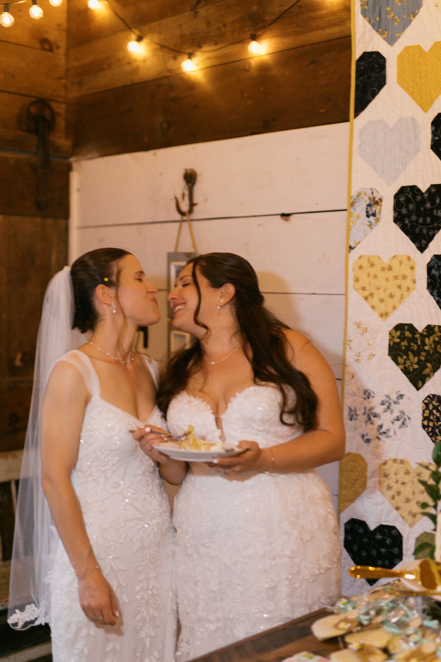 Two brides in white gowns share a tender moment at their wedding reception, holding cake near a decorative heart quilt backdrop.