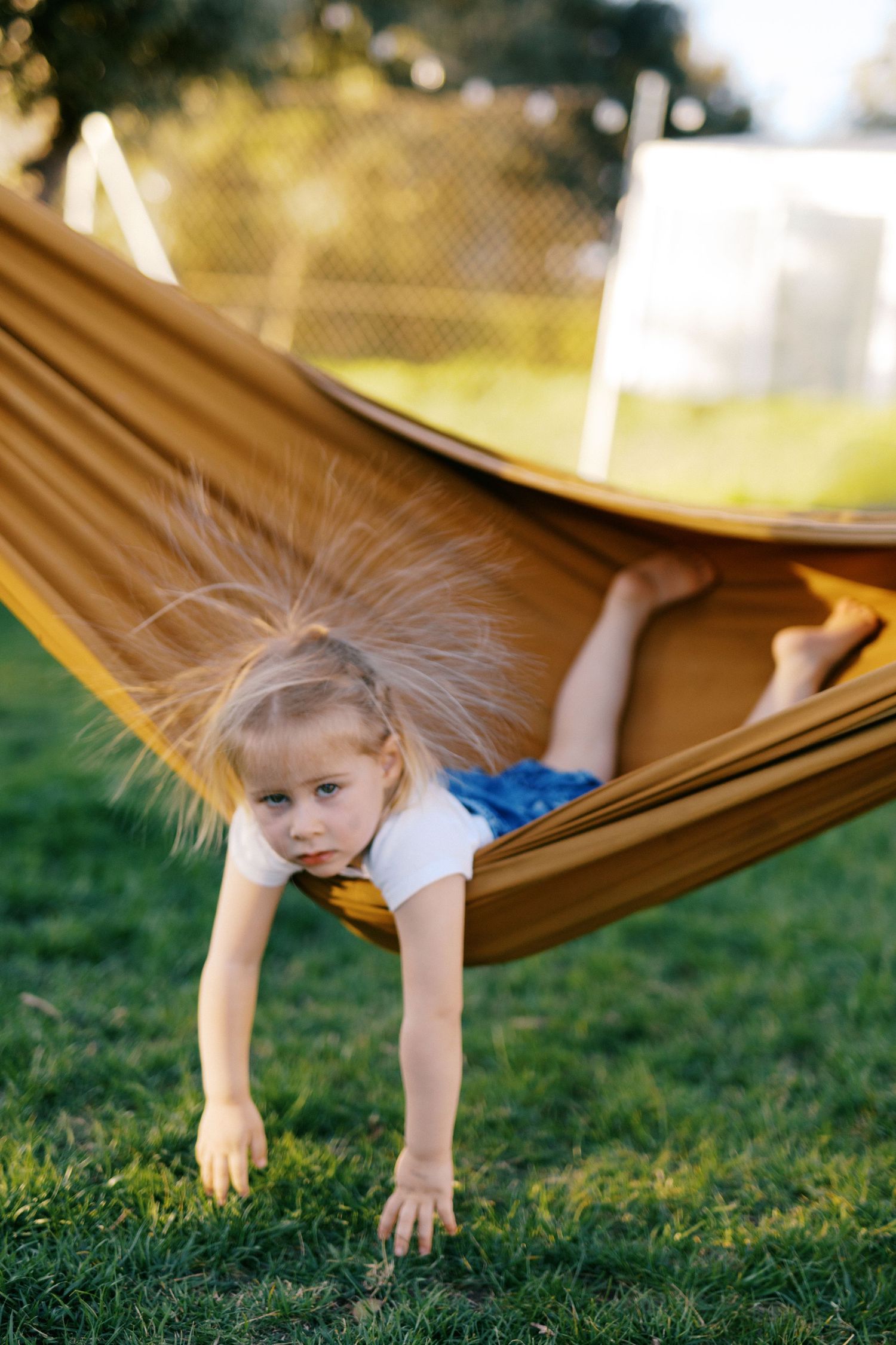 Young blonde girl with static hair lying in a yellow hammock outdoors on green grass.