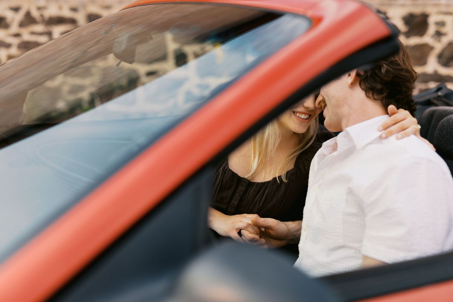 Couple sharing a romantic moment inside a red convertible car, smiling and holding hands.