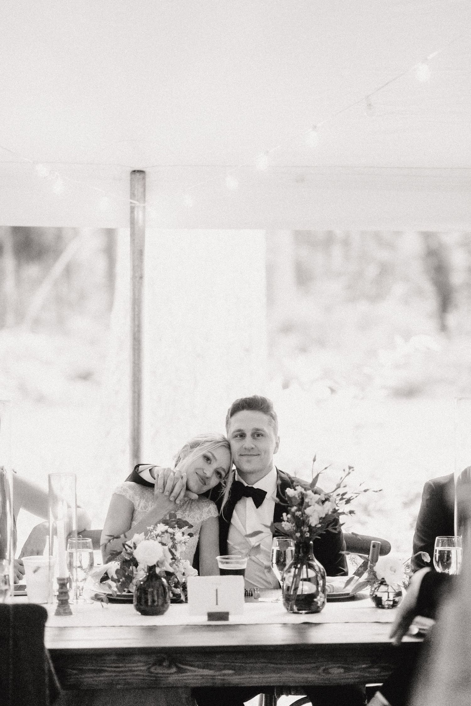 Wedding guests seated at a long rustic wooden table with floral centerpieces at an outdoor reception venue.