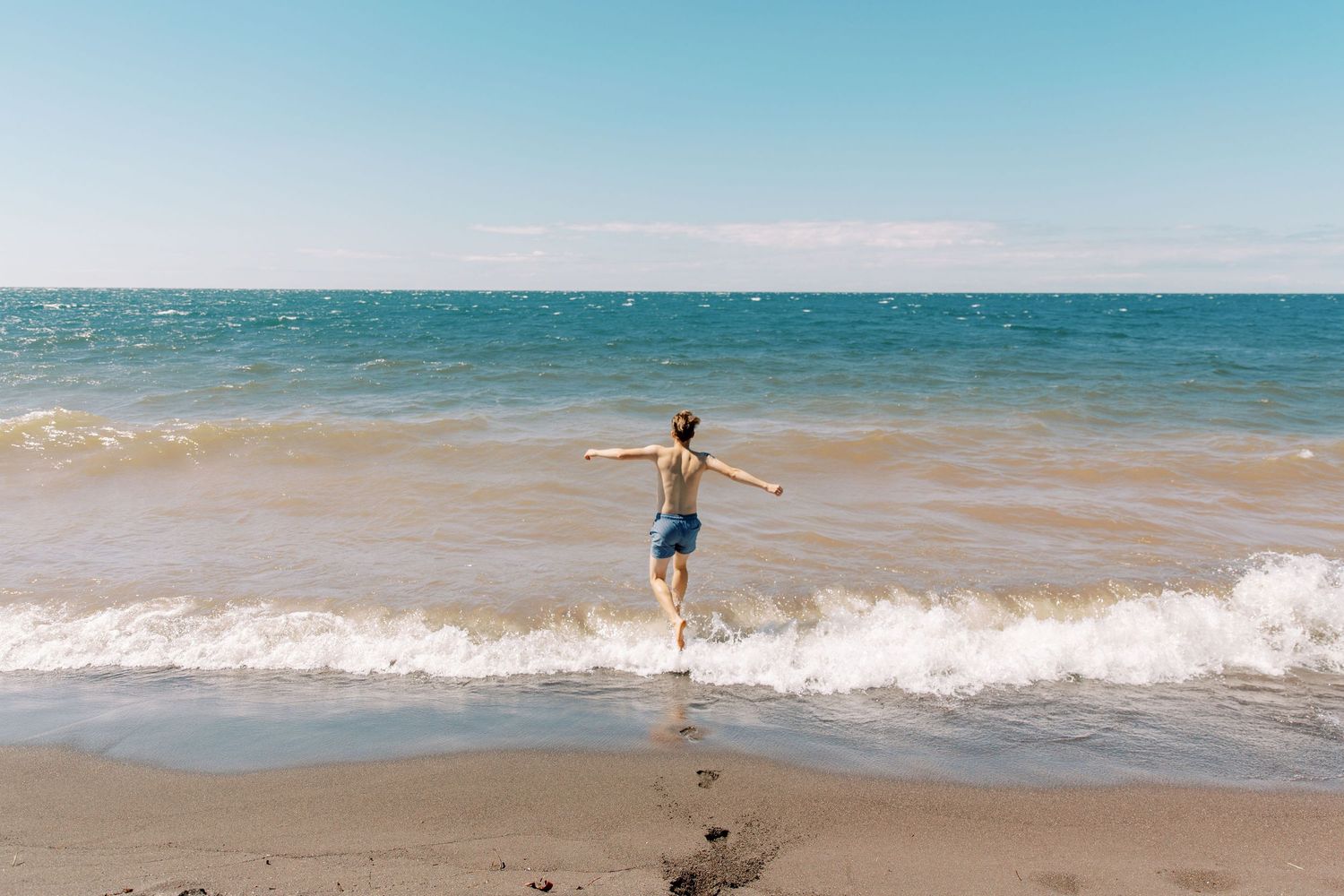 Dylan, Marquette Wedding Photographer, jumps with outstretched arms at the edge of lake waves on a sunny beach with blue water and sky.