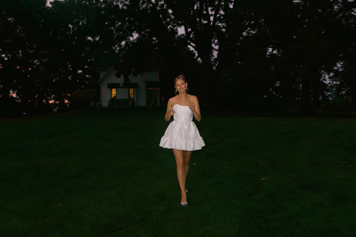 A bride in a white strapless dress walks across a dark lawn at dusk with a house visible in the background.