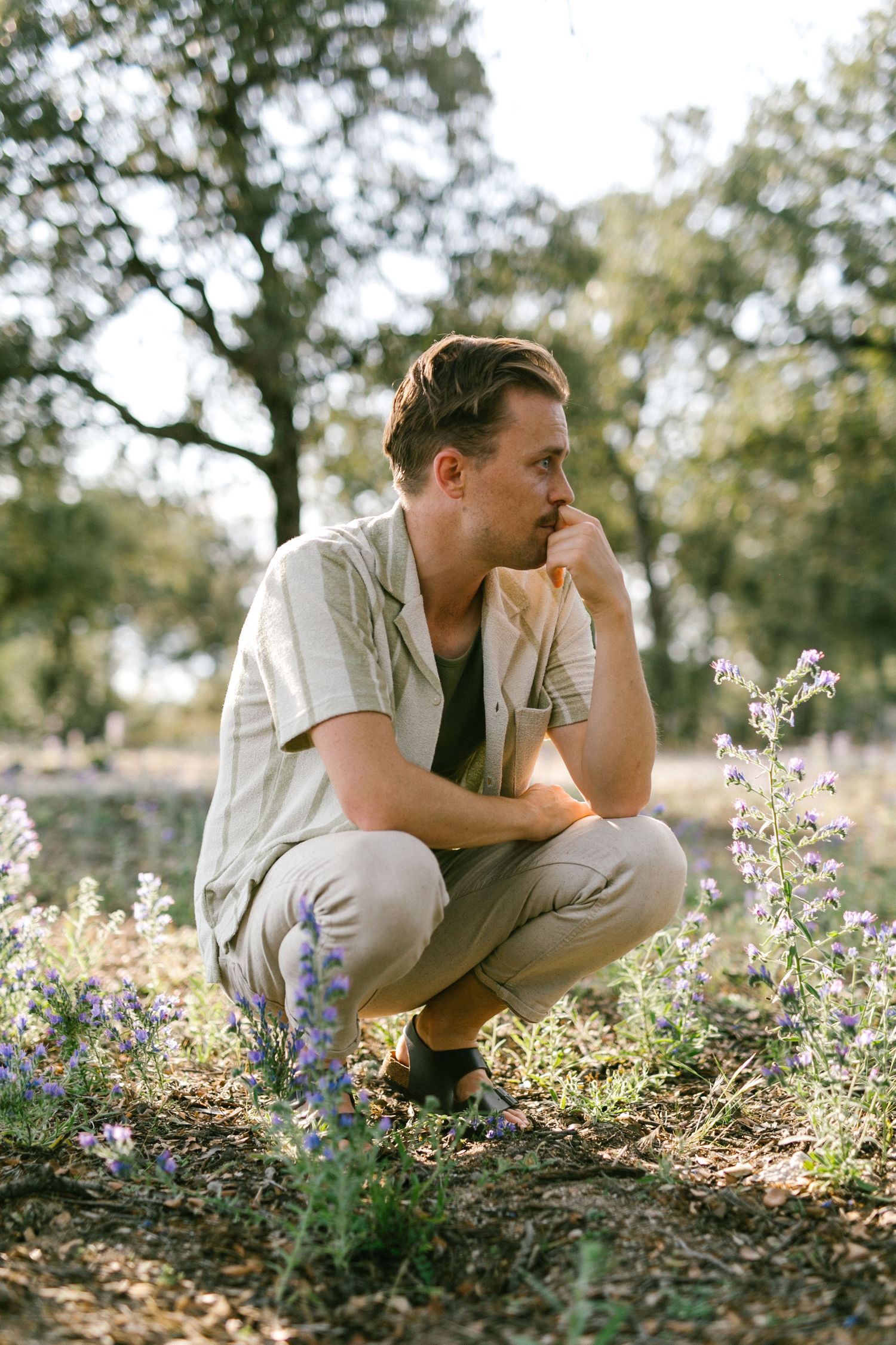 Man in beige outfit crouching among wildflowers in a sunlit outdoor setting.