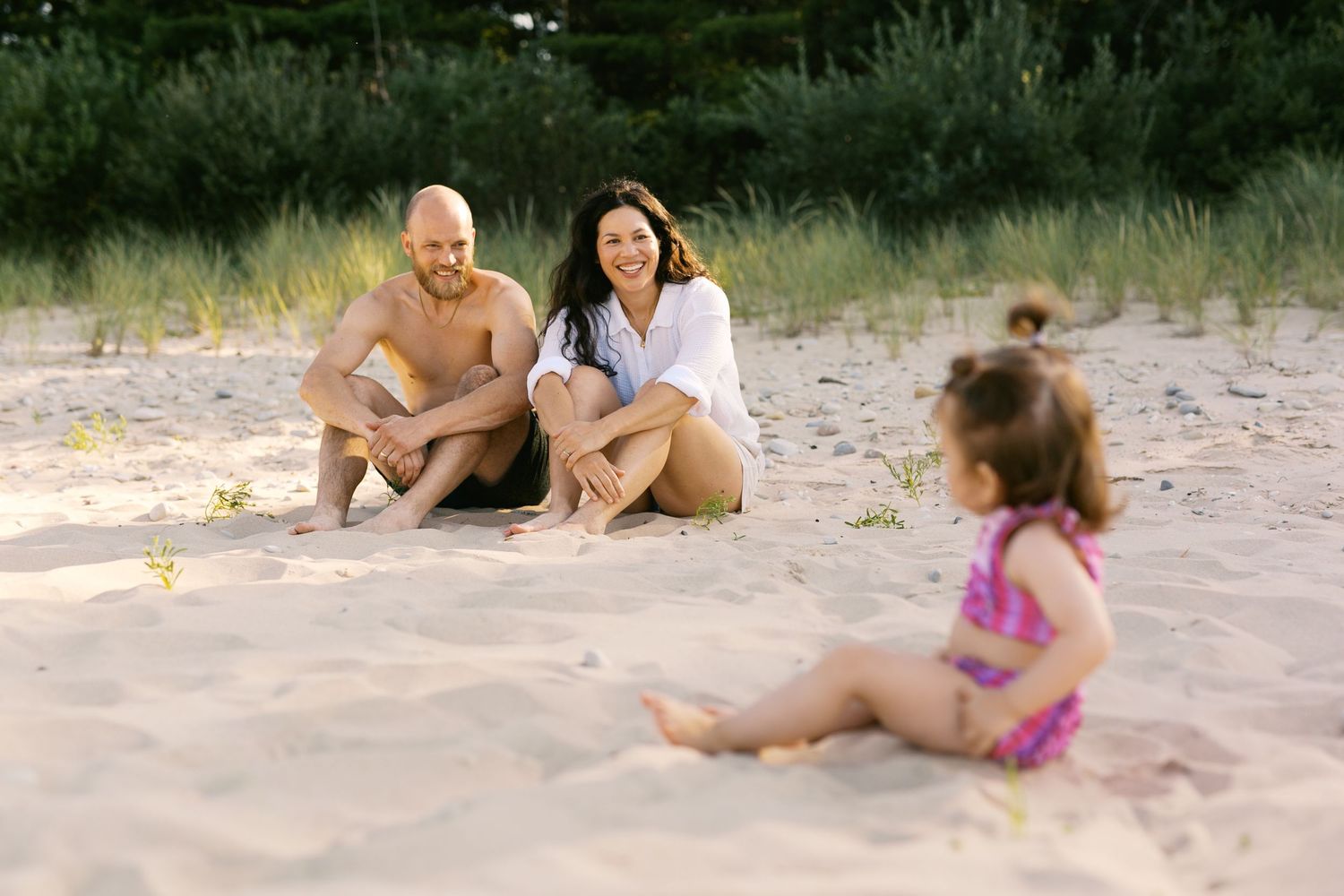 Family sitting on sandy beach, parents smiling at toddler in pink swimsuit during golden hour.