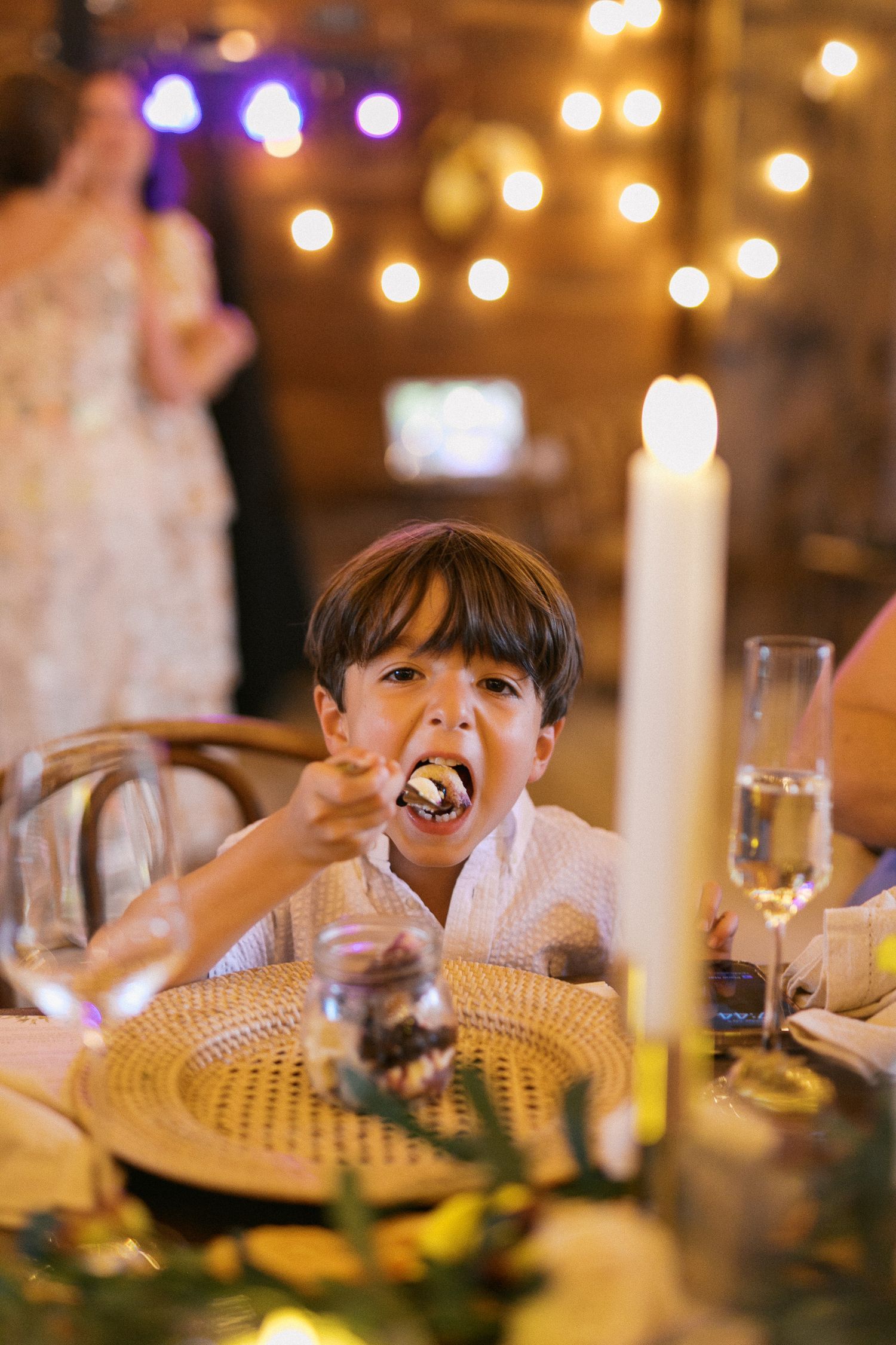 A young boy eating at an elegantly decorated wedding reception table with candles and string lights in the background.